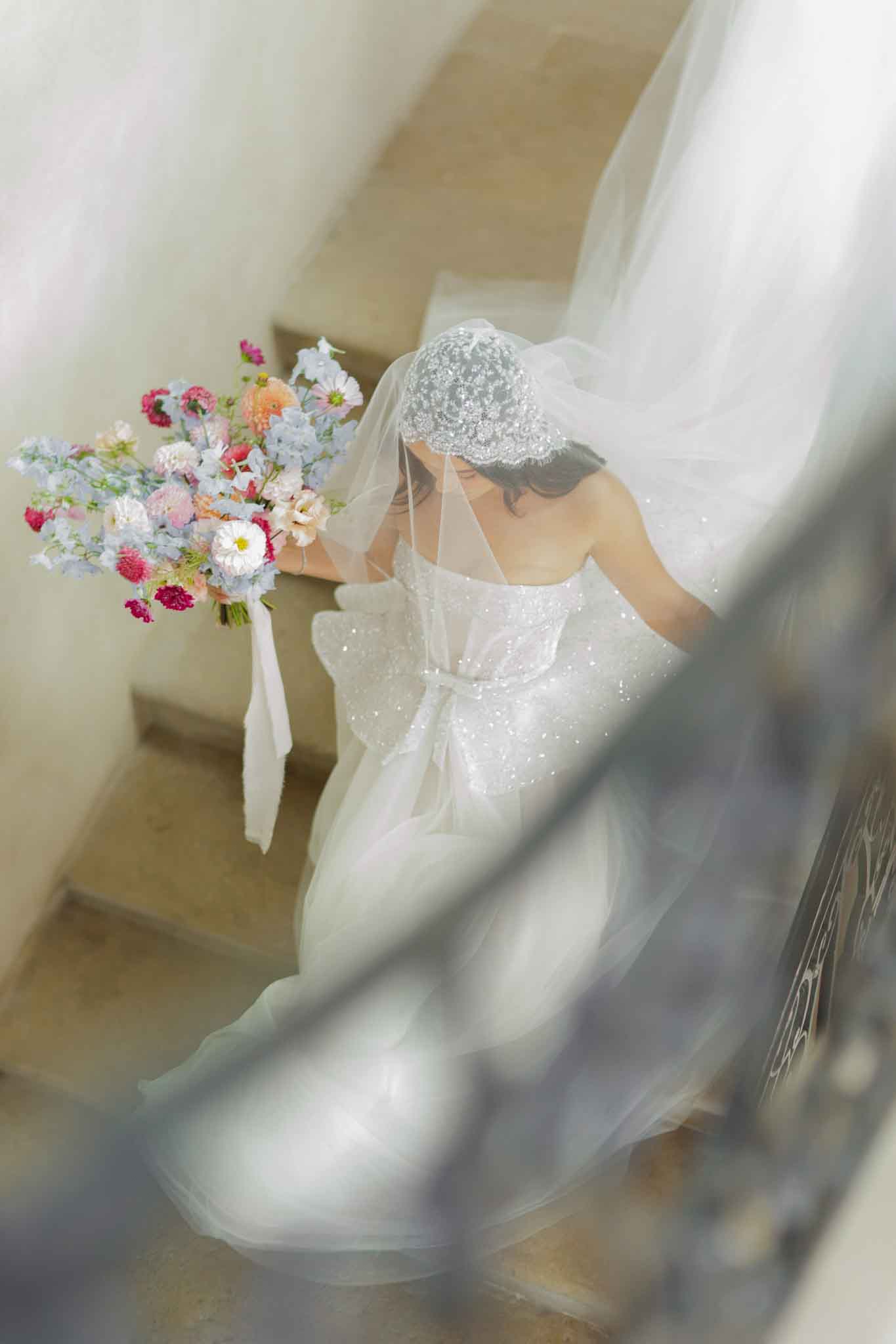 A bride is photographed from above as she descends an interior stone staircase, shot through soft foreground elements including sheer fabric and a wrought-iron railing that frame the composition. She wears a white sparkle-embellished strapless gown with a large bow detail at the back, paired with a pearl and beaded Juliet cap veil with a long tulle train. Her colorful bridal bouquet is a loose, garden-style arrangement featuring dusty blue delphiniums, hot pink and magenta scabiosa, peach ranunculus, soft pink lisianthus, white daisies, and trailing ivory ribbons. The overall styling aesthetic is romantic and fashion-forward, mixing a classic Juliet cap with modern embellishment. The shot is a top-down portrait taken indoors in what appears to be a chateau or manor house interior.