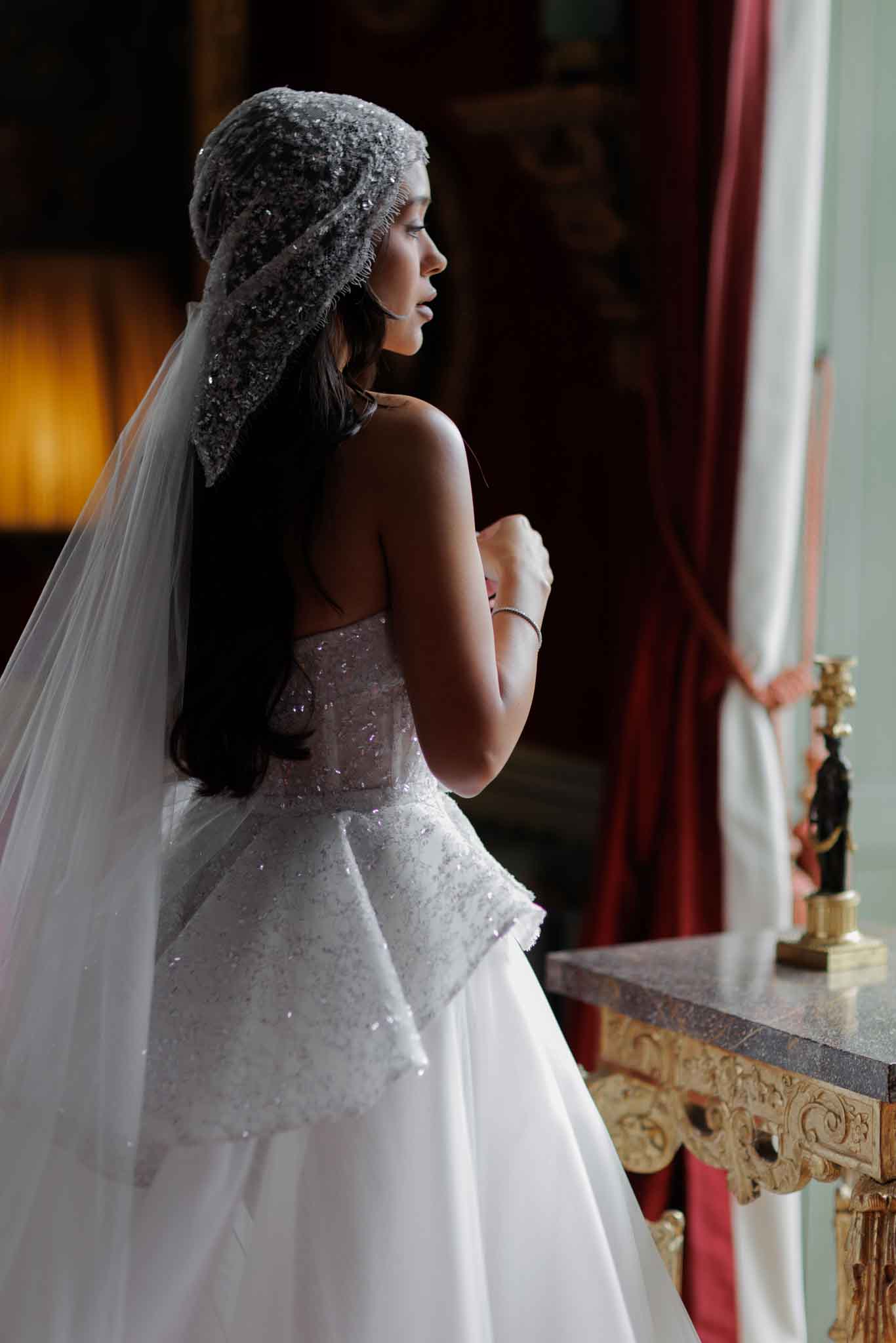 A bridal portrait taken indoors in what appears to be a formal chateau or palace interior, featuring deep red draped curtains, a gilt-legged marble console table, and a warm-toned table lamp visible in the background. The bride stands in three-quarter profile facing a window, wearing a strapless white ball gown with a heavily beaded and structured bodice featuring silver and pale pink embellishment, paired with a flowing tulle skirt. She wears a distinctive heavily beaded and sequined grey cap-style veil that covers the crown of her head and flows into a long white cathedral-length veil, creating a couture-inspired, classic formal aesthetic. The composition is a medium portrait shot with soft natural window light illuminating her profile against the darker ornate interior.