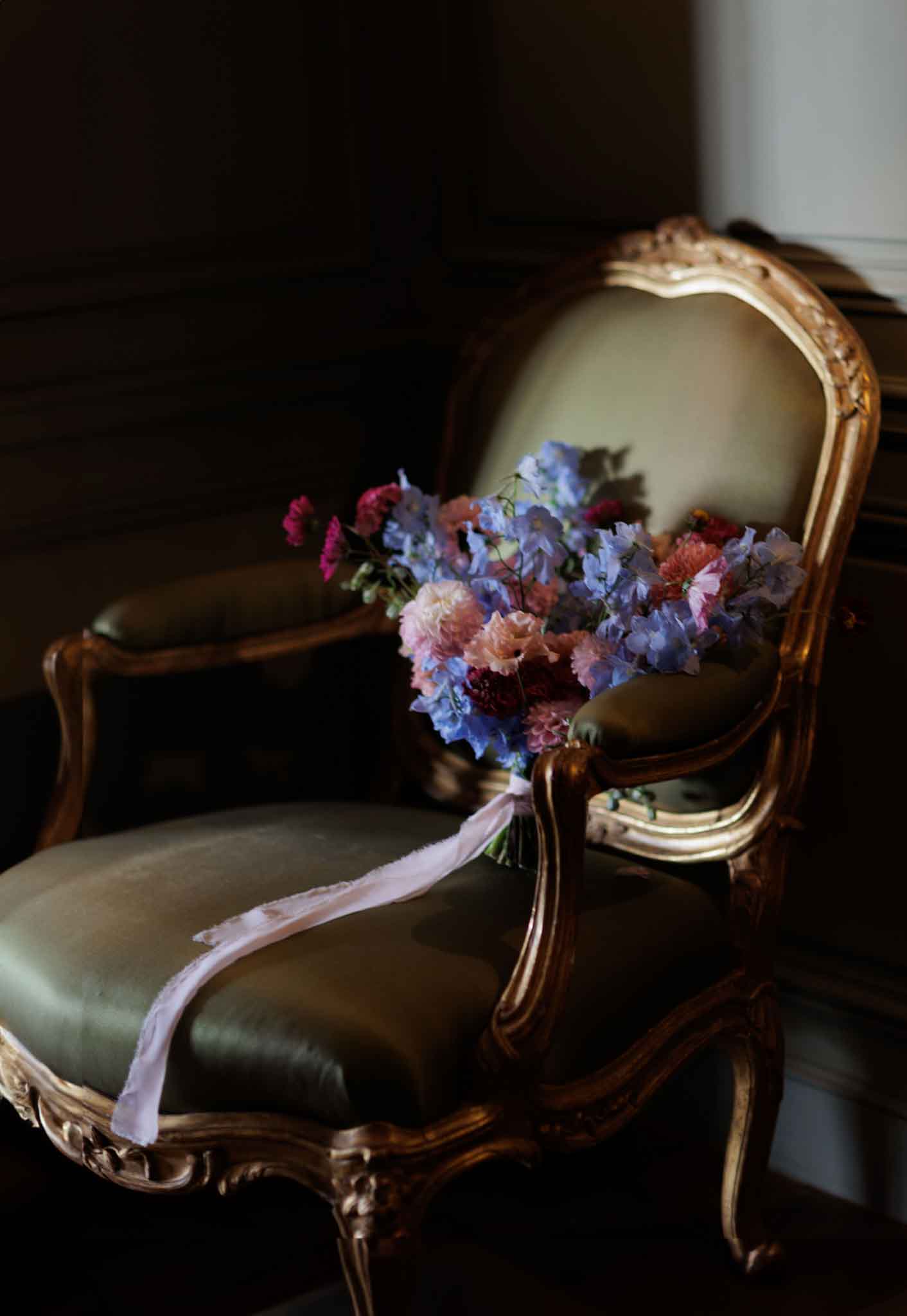 A detail shot of a bridal bouquet resting on the back of a gilded Louis XVI-style armchair upholstered in olive green velvet, set in a dark wood-paneled interior. The bouquet features periwinkle blue delphiniums, magenta and blush sweet peas, dusty pink carnations, and deep burgundy ranunculus, tied with a trailing blush silk ribbon that drapes over the chair seat. The moody, low-key lighting creates strong contrast between the dark background and the vibrant florals. The overall styling is classic and romantic with a jewel-toned floral palette.