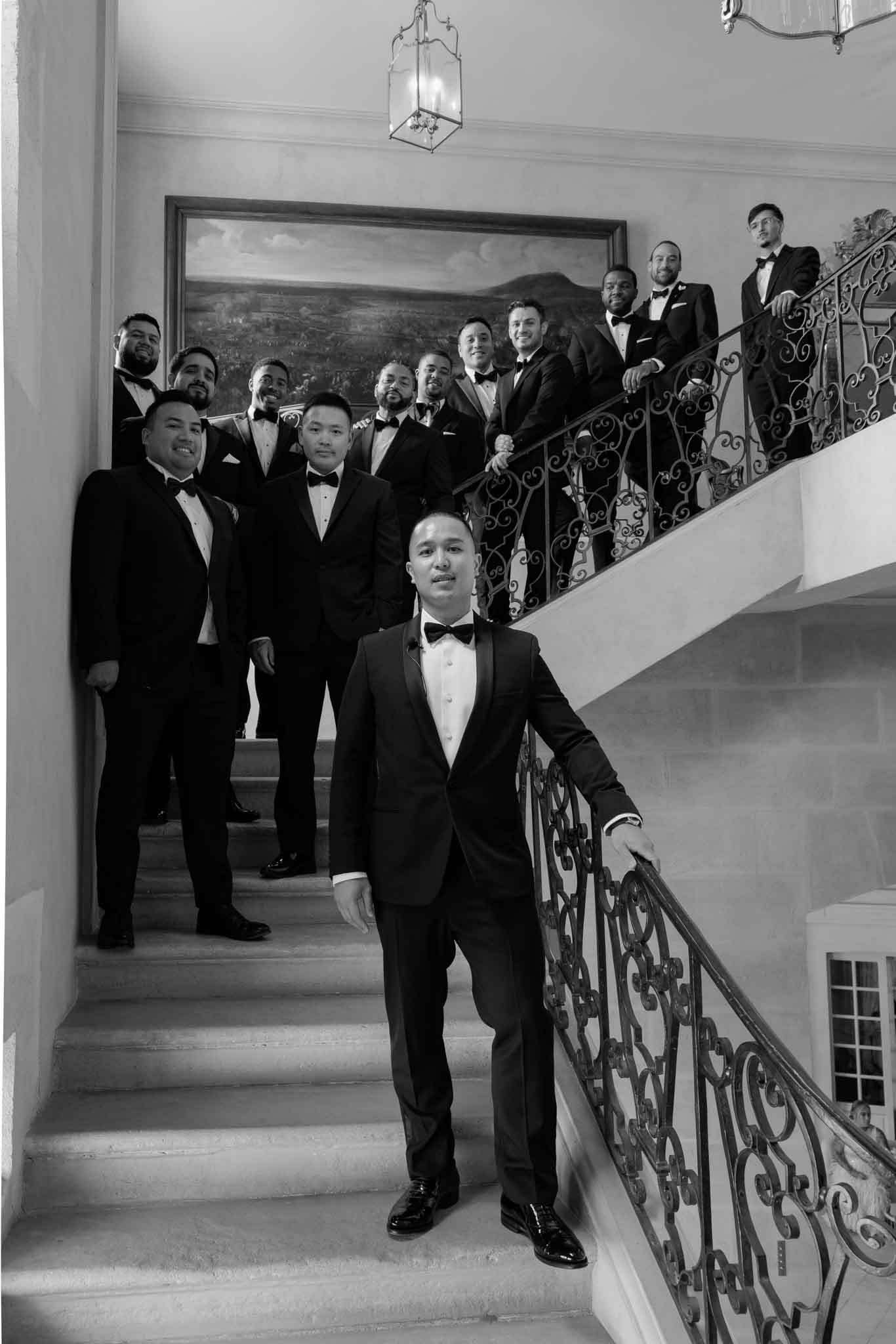 A black-and-white group portrait of a groom and approximately eleven groomsmen posed on a grand interior staircase. The groom stands at the foreground on the lower steps, resting one hand on the ornate wrought-iron banister, while the groomsmen are arranged behind and above him on the staircase. All men are dressed in black tuxedos with bow ties and white dress shirts, styled in a classic formal look. A large framed landscape painting hangs on the wall behind the group, and a glass-paneled pendant lantern is visible at the top of the frame. The high-contrast black-and-white tones emphasize the ironwork details of the banister and the architectural molding of the interior. This is a wide portrait shot taken from a low angle looking up the staircase.