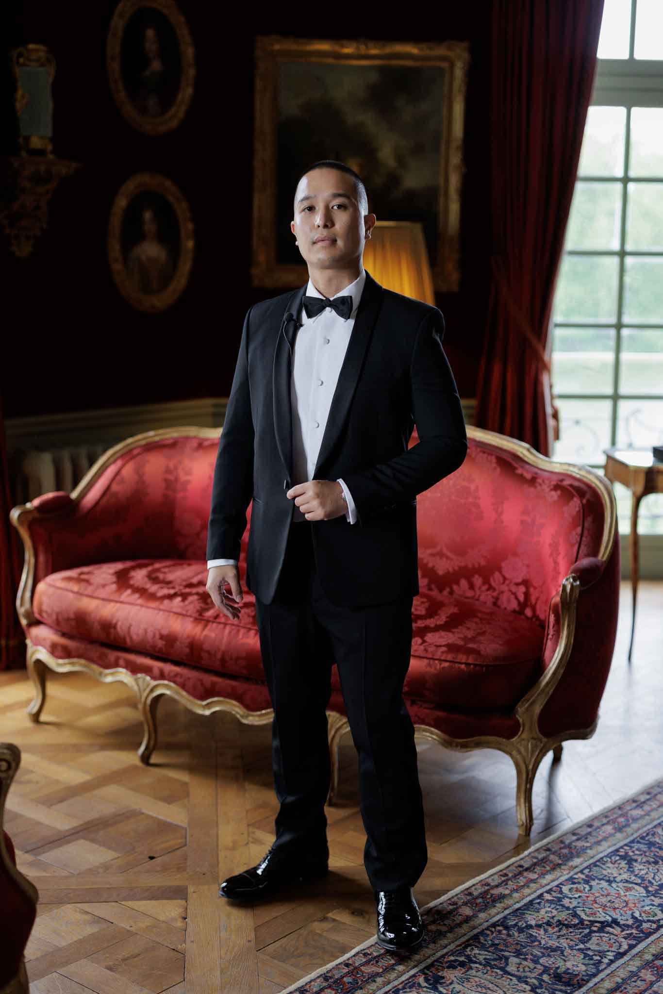A full-length portrait of the groom standing indoors in what appears to be a formal salon or reception room of a French chateau. He is dressed in a black tuxedo with a white dress shirt, black bow tie, and black patent leather shoes, and is adjusting his jacket while looking directly at the camera. The room features a red damask Louis XV-style canapé sofa with gold gilt frame, dark walls hung with ornate oval-framed portrait paintings, deep red curtains, parquet herringbone flooring, a patterned area rug, and a lit table lamp in the background. The overall decor palette is deep red, black, and gold, consistent with a classic French chateau interior.