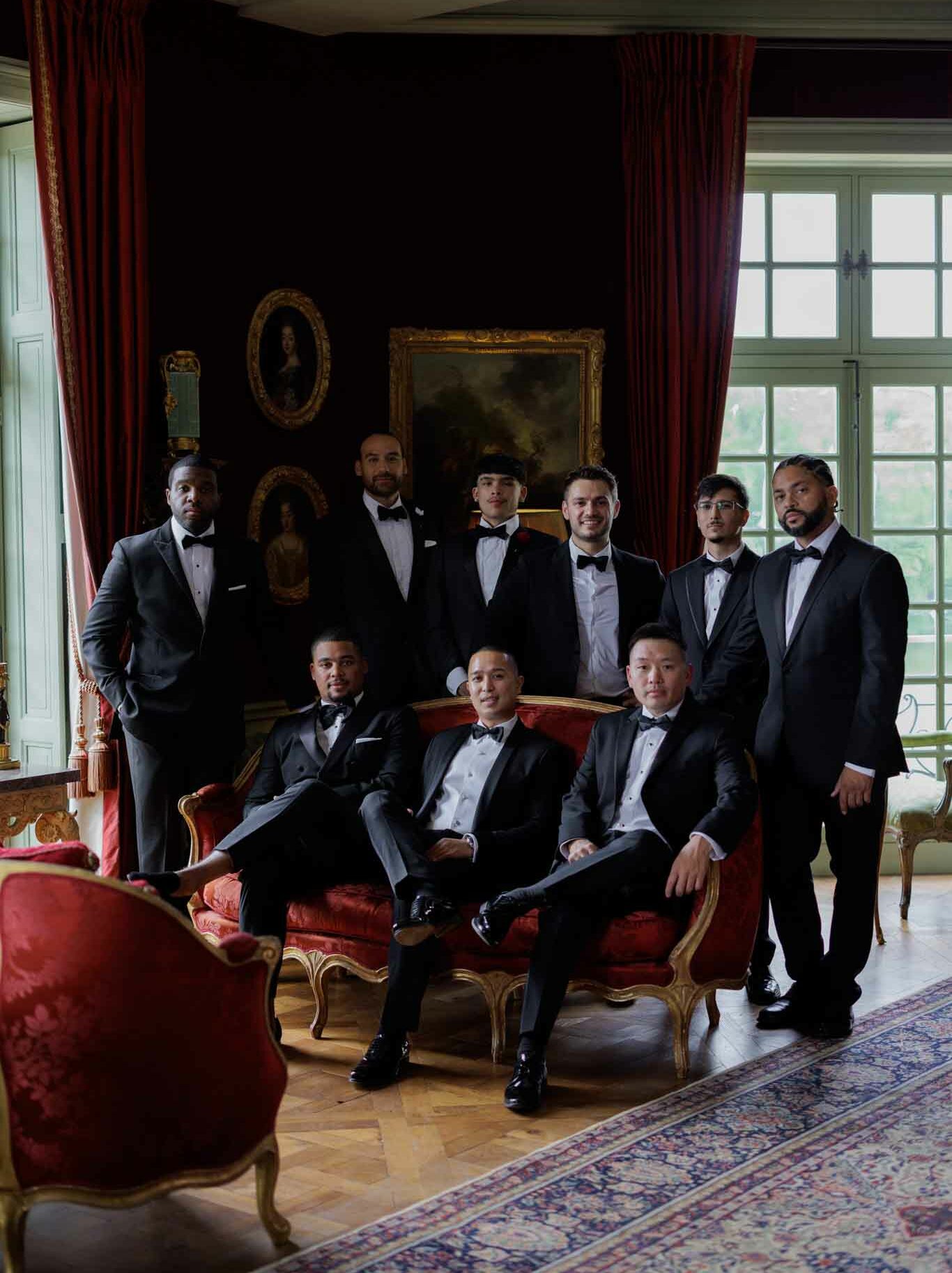 A groomsmen portrait taken indoors in what appears to be a formal salon of a French chateau, featuring nine men in black tuxedos with white dress shirts and black bow ties. Three men are seated on a gold-framed red damask settee, while six stand behind and to the sides, arranged in a structured group pose. The room features dark walls, floor-length deep red velvet drapes, gold-framed oil paintings including a large landscape and an oval portrait, parquet wood flooring, and a patterned area rug, all contributing to a classic, formal interior setting. The lighting is moody and directional, with natural light entering from tall multi-pane windows on the right. This is a medium wide-angle portrait shot.