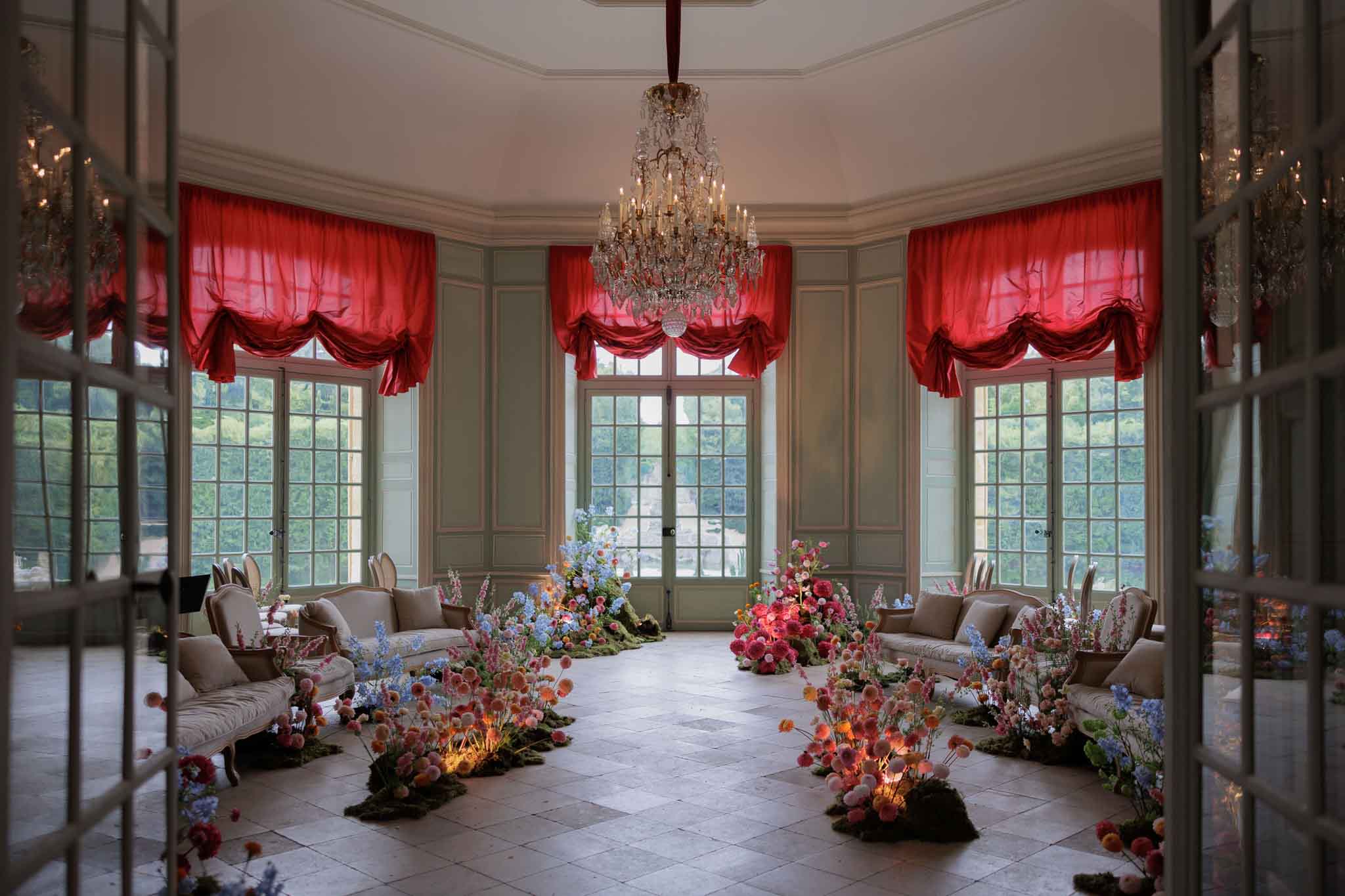 An indoor ceremony or cocktail space set up inside a classic French chateau salon, photographed as a wide interior shot through open mirrored double doors. The octagonal room features sage green painted paneling, tall multi-pane French doors with crimson red draped balloon-style curtains, and a large crystal chandelier hanging from the center of the ceiling. The stone-tiled floor is lined on both sides with cream-upholstered Louis XVI-style sofas and settees. Elaborate floor-level floral installations run along the perimeter and down the center aisle, composed of clustered arrangements in coral, hot pink, peach, lavender blue, and soft orange — including what appear to be dahlias, delphiniums, poppies, and alliums set among beds of moss, with small warm-toned uplights illuminating the blooms from below. No people are present; the space is empty and styled for an event. Potential venue feature image.