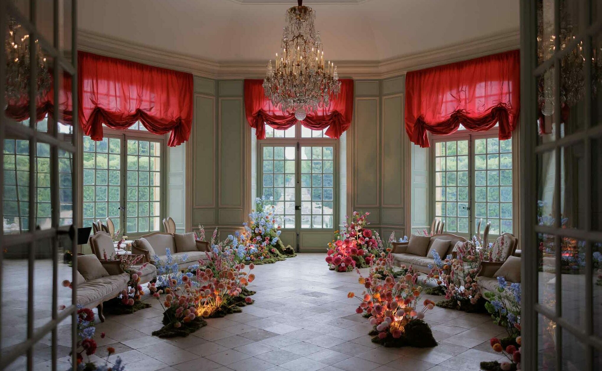 A wide shot of an indoor cocktail hour or reception lounge space inside a French chateau-style ballroom, photographed through open glass-paned doors. The room features sage green wood-paneled walls, tall multi-pane French windows dressed with crimson red draped silk swag curtains, and a large crystal chandelier with candle-style lights hanging from a white ceiling with crown molding. The floor is laid in large stone tiles in a diagonal pattern. Several cream-upholstered Louis XVI-style curved sofas and armchairs are arranged symmetrically around the perimeter of the room. The dominant decorative feature is a series of dramatic garden-style floral installations placed directly on the floor throughout the space — large, low, moss-based arrangements bursting with pink dahlias, coral and peach ranunculus, blue delphiniums, pink foxglove, and assorted wildflower-style blooms. Small warm accent lights are placed within the floral clusters, illuminating them from below with an amber glow. The overall decor style is maximalist and romantic, combining classical French interior architecture with loose, organic floral design. No people are present in the image. Potential venue feature image.