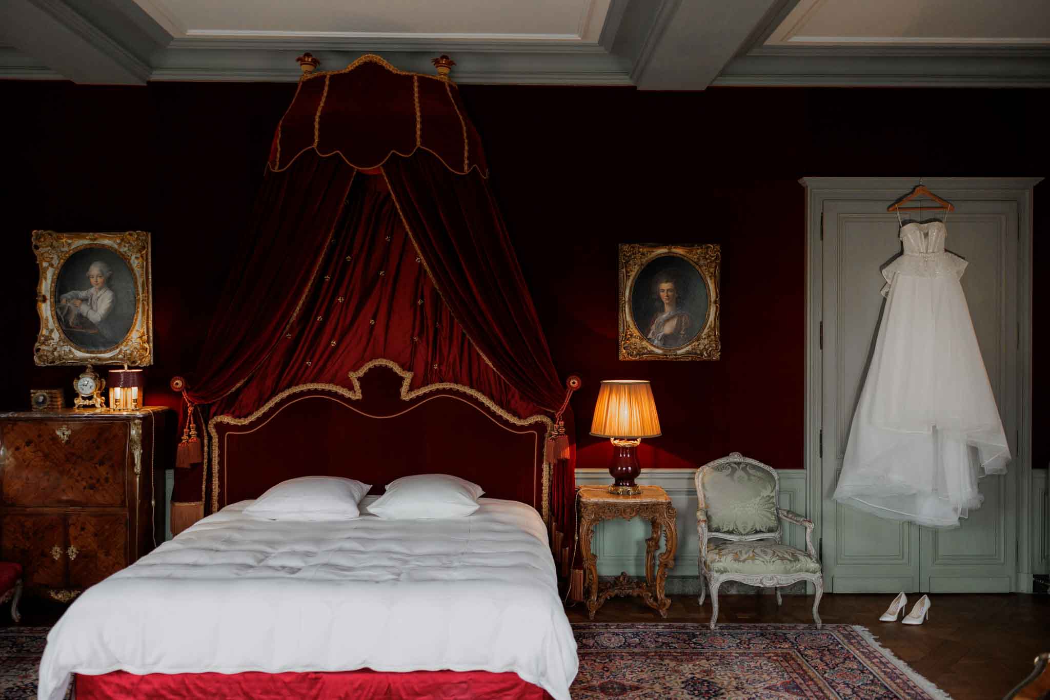 A bridal getting-ready detail shot taken in an ornate chateau bedroom, with no people present. The white strapless wedding dress with a full tulle skirt hangs from a wooden hanger on the door of a sage green armoire at the right of the frame, with a pair of white satin heels placed on the floor beneath it. The room features deep crimson walls, a grand canopied bed with a burgundy velvet and gold-trimmed headboard and matching draped corona, two gilt-framed oil portrait paintings, a gilded Rococo-style bedside table with a warm amber lamp, an antique walnut chest of drawers, a sage green Louis XV-style armchair with tapestry upholstery, and a Persian rug on the parquet floor. The overall decor style is classic French aristocratic with rich, warm tones. Wide-angle interior shot capturing the full room composition.