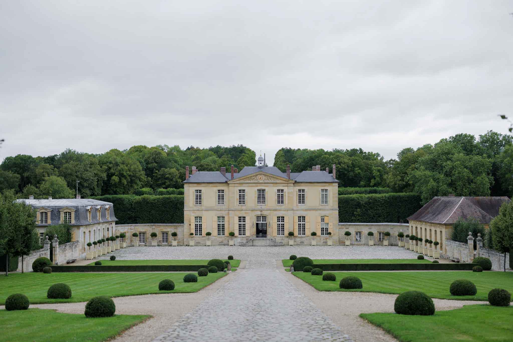 Wide exterior shot of a French classical chateau viewed from the end of a formal gravel approach path lined with symmetrically arranged clipped box ball topiaries set into gravel and lawn parterres. The main building is a two-story pale limestone structure with a slate mansard roof, multiple tall windows, a central pediment, and a small cupola. Curved colonnaded wings extend on either side, connecting to flanking outbuildings, with potted standard topiary trees lining the forecourt walls. No people are visible; this appears to be an establishing shot of the venue grounds and architecture. Potential venue feature image.