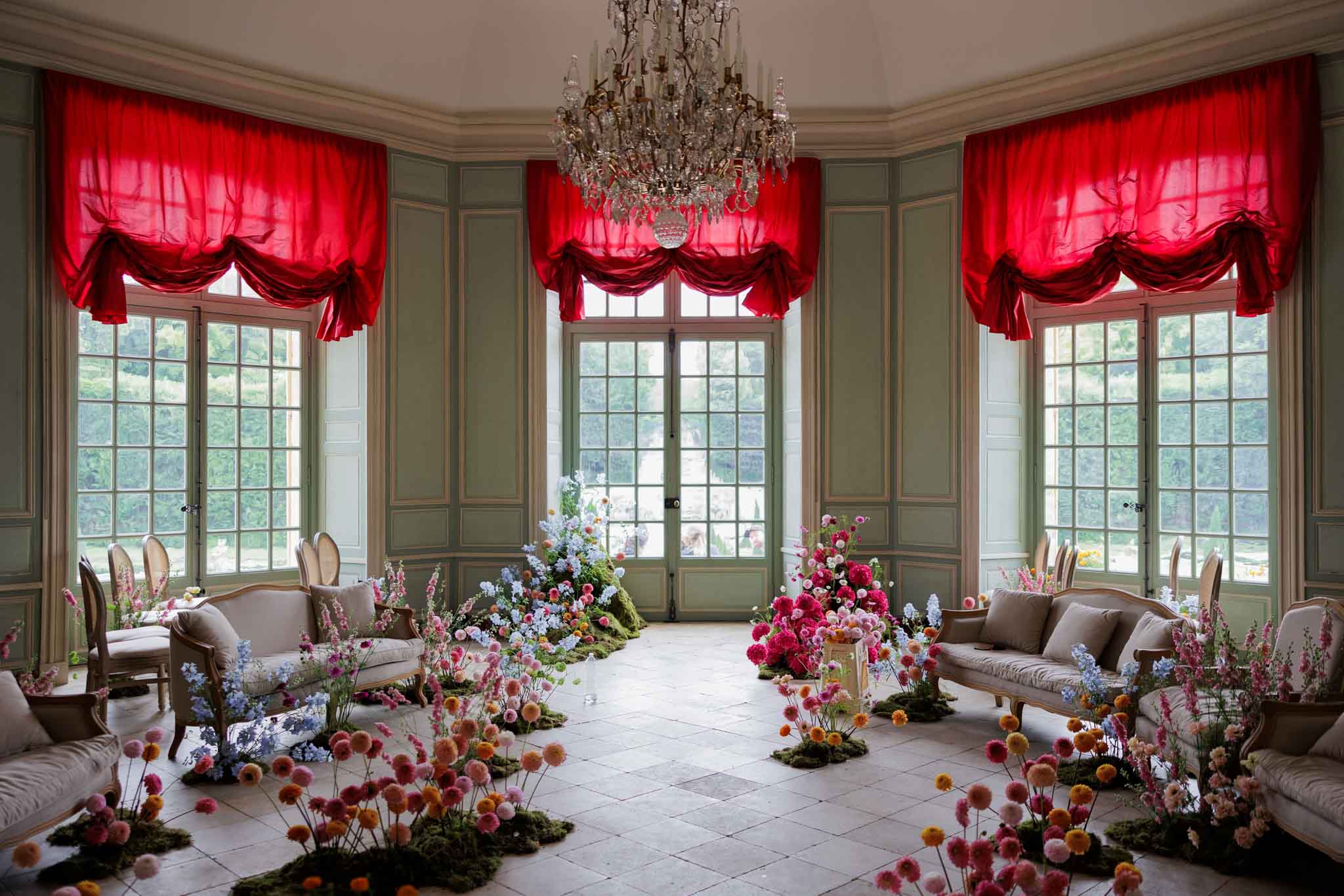 An indoor ceremony or cocktail space set up inside what appears to be a French chateau salon, featuring sage-green paneled walls, three large floor-to-ceiling French windows with bold crimson red draped curtains, and a crystal chandelier overhead. The stone-tiled floor is lined on both sides with cream-upholstered Louis XVI-style sofas and armchairs, arranged to create a central aisle. Elaborate floral installations are scattered across the floor in moss-covered clusters, featuring hot pink dahlias, light blue delphinium, soft pink and peach globe amaranth (gomphrena), orange ranunculus, and white blooms, creating a garden-floor effect that extends toward the central doorway. The overall decor style is maximalist and garden-inspired, with a rich contrast between the crimson drapery and the abundant pastel-to-vivid floral palette. Wide symmetrical shot from the entrance of the room. Potential venue feature image.