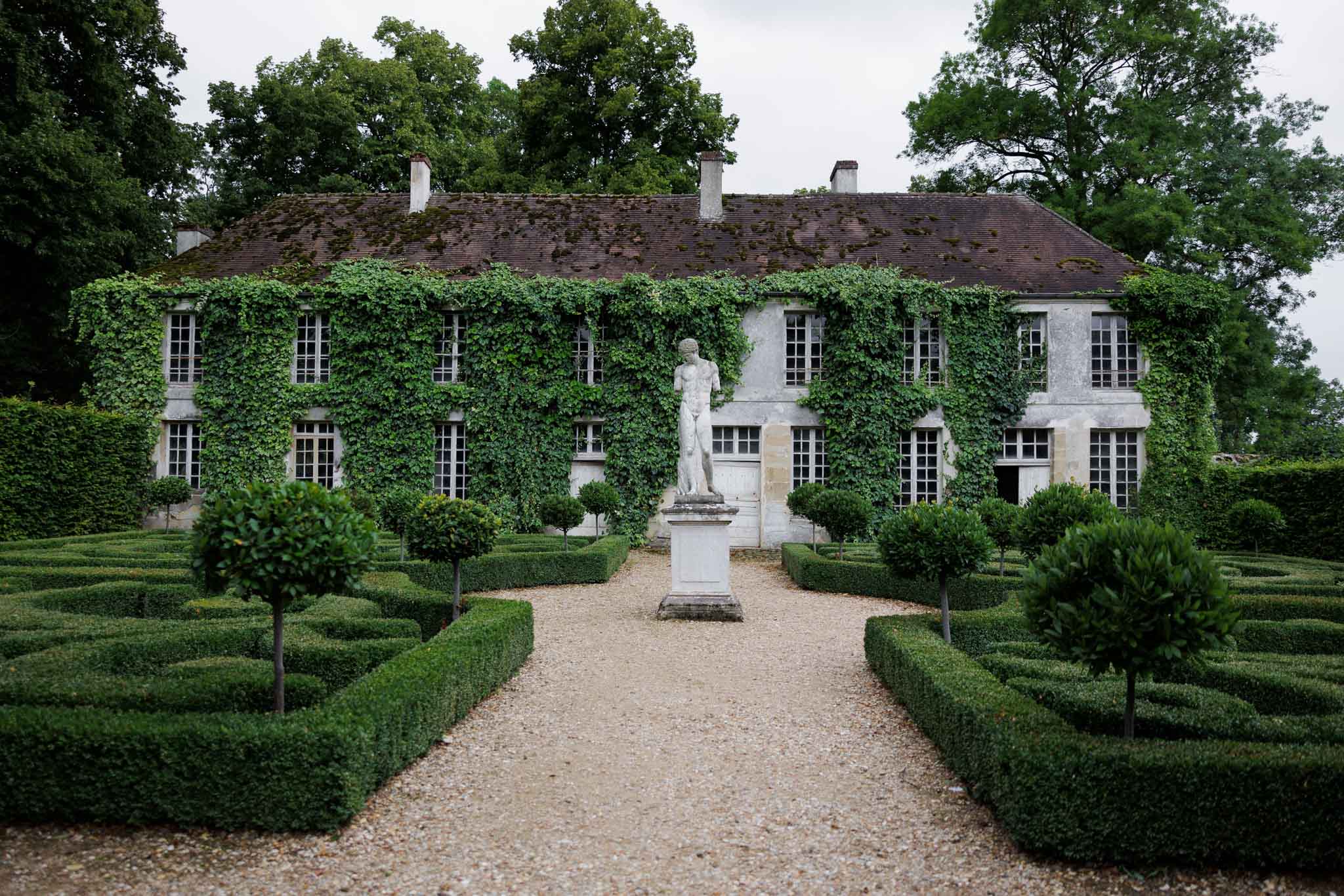 Wide shot of the exterior facade of a French country manor or château, with no wedding party or guests visible. The two-storey stone building is heavily covered in climbing ivy, with rows of white-framed multi-pane windows across both floors. In the foreground, a formal French parterre garden features precisely clipped low box hedges arranged in geometric patterns, standard lollipop-shaped topiary trees lining a central gravel path, and a white classical figurative statue on a stone pedestal positioned in the middle ground directly in front of the building's entrance. The overall palette is grey stone, white trim, and deep green topiary. Potential venue feature image.