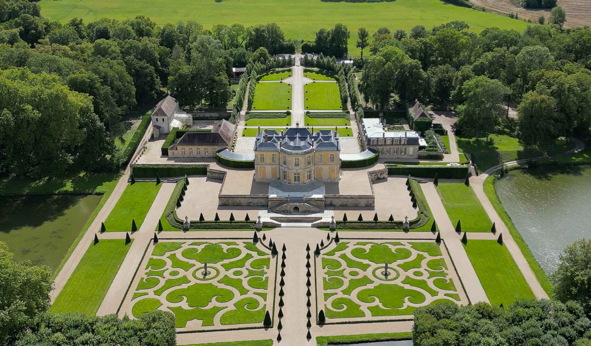 Aerial wide shot of a French classical chateau with a pale yellow stone facade and dark mansard roof, surrounded by formally designed grounds in the French formal garden style. The front grounds feature two large symmetrical parterre gardens with ornate scrollwork hedging patterns, clipped conical topiary, and central fountain features, all bordered by gravel pathways. Behind the chateau, a long central allée flanked by trimmed hedgerows and additional formal garden sections extends toward open countryside, with outbuildings and a moat or river visible on either side of the property. No people or wedding-specific decor are visible in this image. Potential venue feature image.