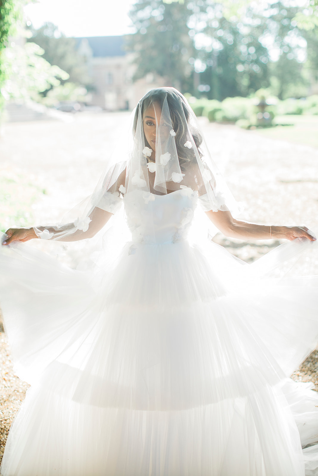 A bridal portrait taken outdoors on a gravel path, with a chateau building and manicured garden visible in the softly blurred background. The bride is facing the camera while spreading her voluminous white tulle ball gown skirt wide with both arms outstretched, and a two-tier veil adorned with scattered white 3D floral appliqués drapes over her face and shoulders. She wears a structured sweetheart-neckline bodice and gold statement earrings, with loose wavy hair beneath the veil. The image is shot in bright, high-key natural light with a slightly overexposed quality, giving the white tulle layers a luminous, airy appearance, captured in a medium full-length portrait composition.