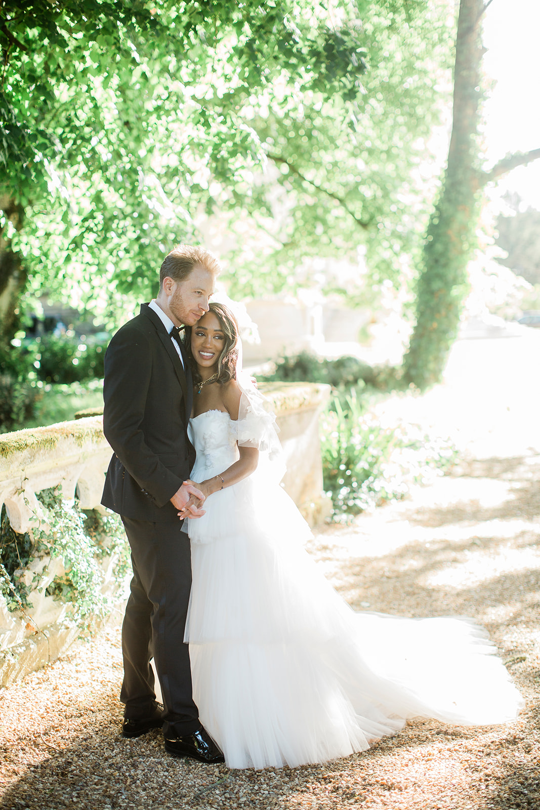 A couple portrait taken outdoors on a gravel path beside a stone balustrade, shot in bright natural light with a soft, airy feel. The bride wears an off-the-shoulder white tulle ballgown with tiered layers, a cathedral-length veil, and a fitted bodice, while the groom is dressed in a black tuxedo with a black bow tie and patent leather shoes. The groom stands behind the bride, leaning his cheek close to hers as she smiles toward the camera, their hands clasped together in front of them. The composition is a full-length portrait with the couple centered in the frame.