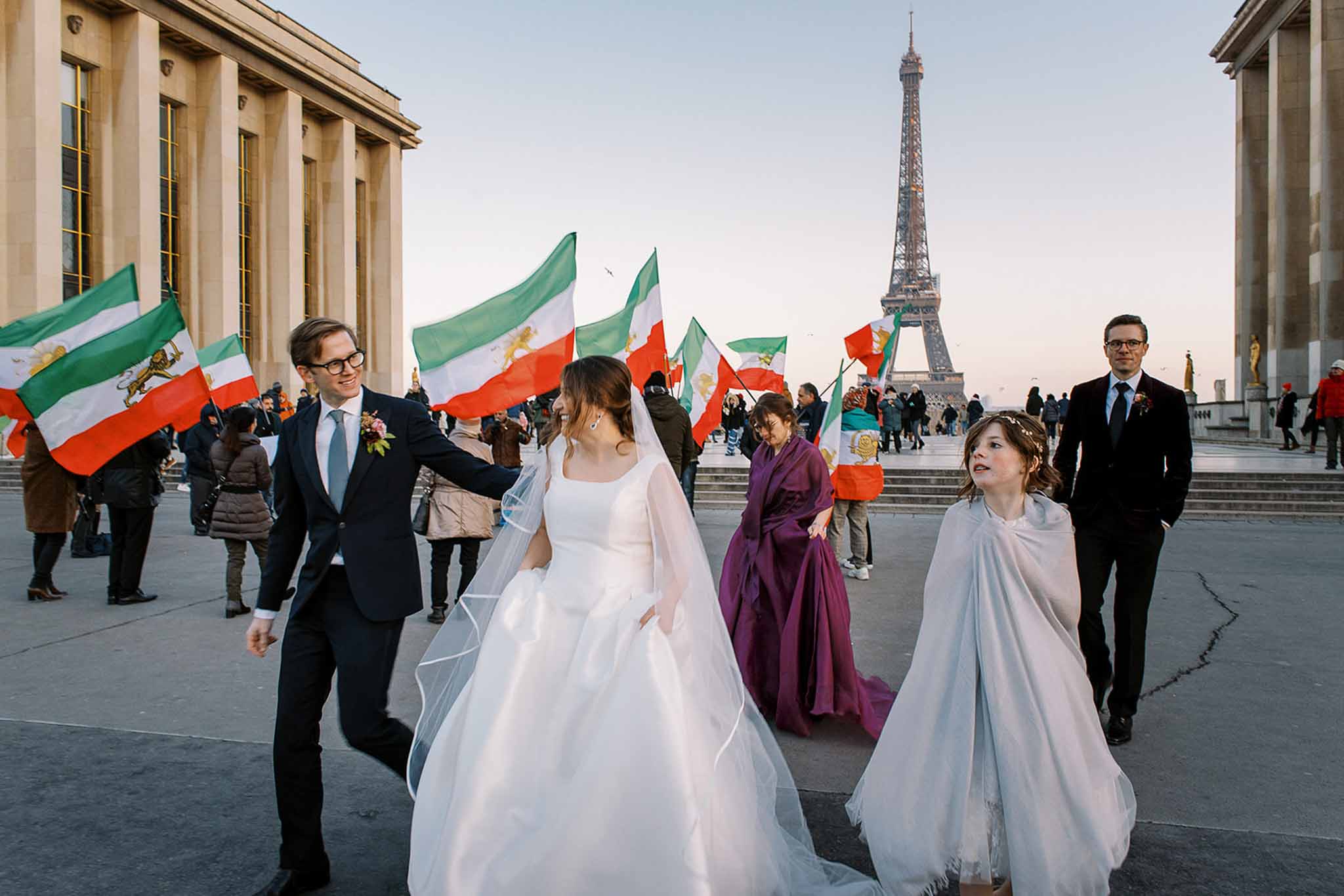 A wedding party walks across the Trocadéro esplanade in Paris with the Eiffel Tower centered in the background. The bride wears a white long-sleeve ball gown with a cathedral-length veil, and the groom is dressed in a navy suit with a light blue tie and a small floral boutonnière. A young flower girl wrapped in a light grey shawl and wearing a floral crown walks alongside them, while a woman in a deep plum gown and a man in a dark suit follow behind. A large public demonstration is taking place simultaneously in the background, with dozens of people waving Iranian imperial lion-and-sun flags across the plaza. The wide shot captures the unplanned, candid energy of the moment, with the wedding party appearing to navigate through the crowd mid-procession.