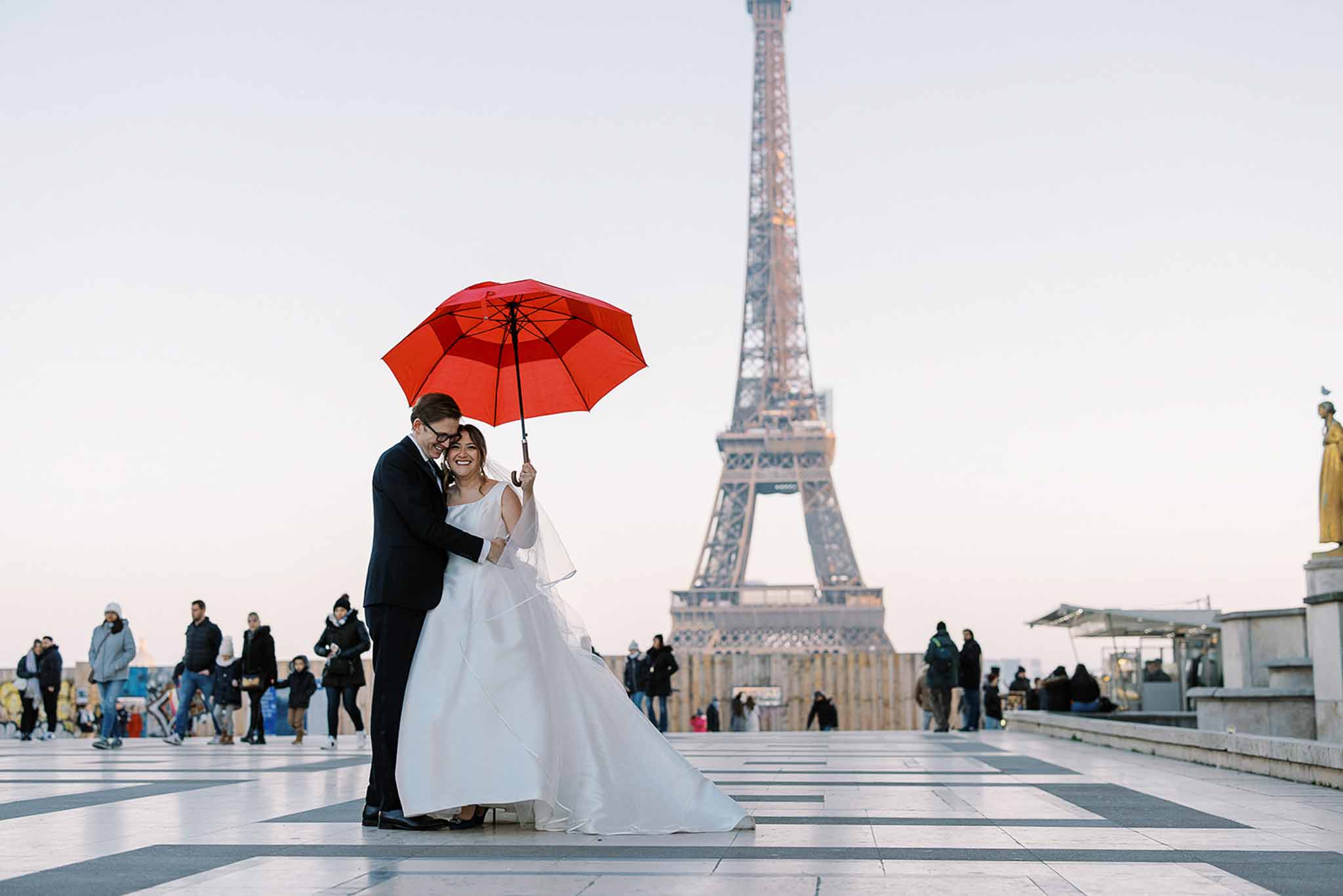 A couple pose for a wedding portrait on the Trocadéro esplanade in Paris, with the Eiffel Tower visible in the background. The bride wears a full-skirted white ballgown with a cathedral-length train and a fingertip veil, while the groom wears a classic black tuxedo with a bow tie and glasses. The bride holds a bold red umbrella overhead, creating a striking color contrast against her white gown, and the couple laugh together as passersby in winter coats move around them. The composition is a wide shot taken at ground level, placing the couple centrally in the foreground with the Eiffel Tower softly out of focus behind them.