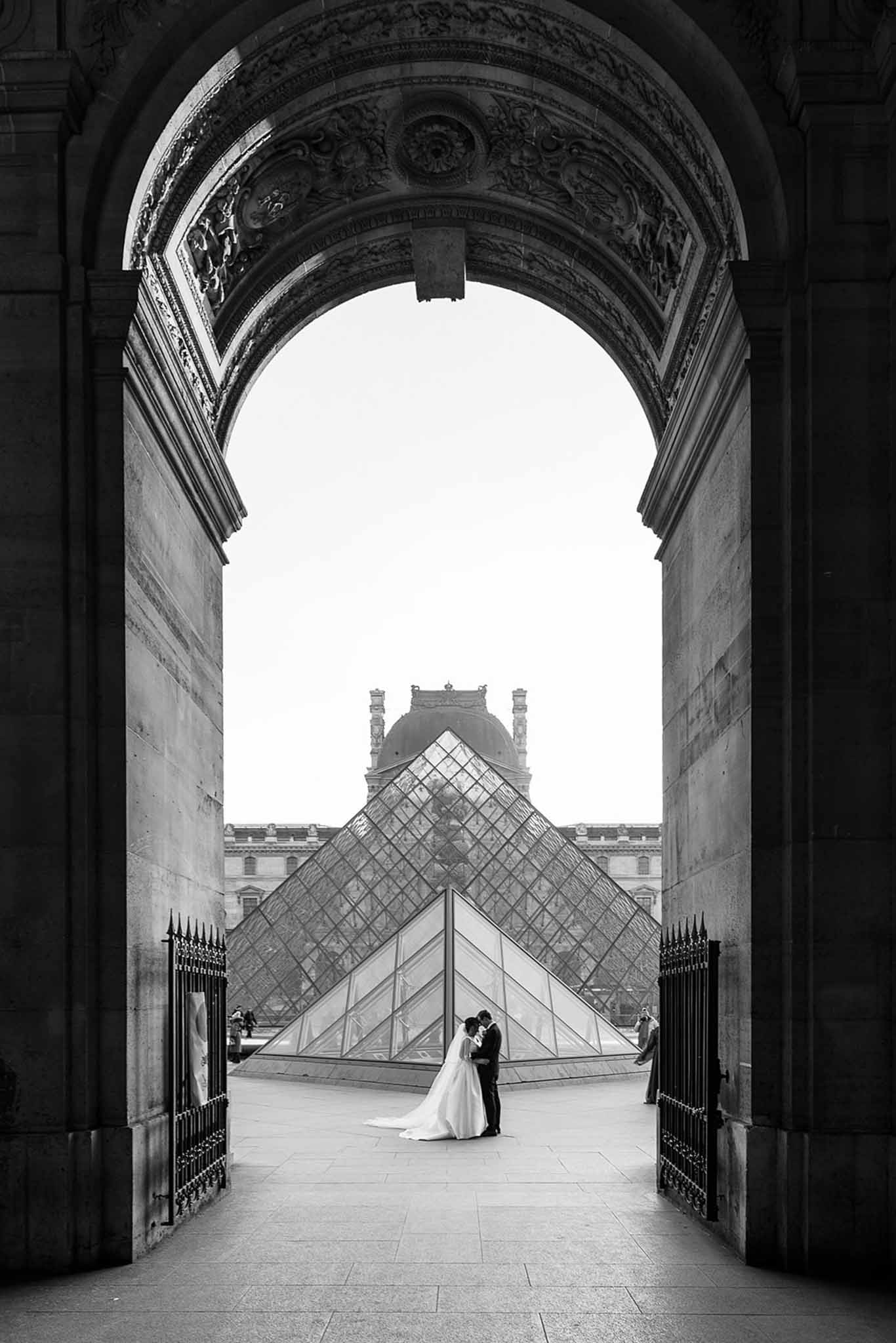 This black-and-white couple portrait is shot from within a large ornate archway of the Louvre museum in Paris, framing the bride and groom as small figures standing close together in front of the iconic glass pyramid in the courtyard beyond. The image has strong contrast with deep dark tones in the carved stonework of the arch and bright highlights on the open sky and pyramid structure. The bride wears a full ballgown with a long train visible on the ground, while the groom is dressed in a dark suit; the two are facing each other in an intimate pose. The composition is a wide architectural shot that uses the arch as a natural frame, placing the couple at the geometric apex of the pyramid, with iron gates flanking them on either side and the classical Louvre palace wings visible in the background.