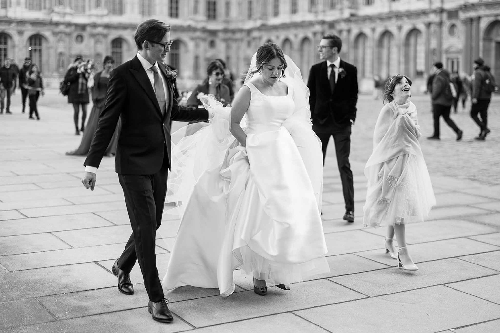 A black-and-white candid portrait of a wedding party walking across a large paved courtyard of a classical Haussmann-style building, identifiable as the Cour Carrée of the Louvre in Paris. The bride wears a full-skirted, sleeveless ball gown with a square neckline and a long veil, and the groom — in a dark suit with a light tie and boutonnière — walks beside her helping to manage the veil in the wind. To their right, a female guest in a short lace dress and white wrap shawl laughs openly, while a second man in a dark suit and tie walks behind them. Passersby and tourists are visible in the background, adding to the candid, documentary feel of the shot. The image has strong contrast with bright highlights on the bride's gown and mid-toned shadows across the courtyard paving.