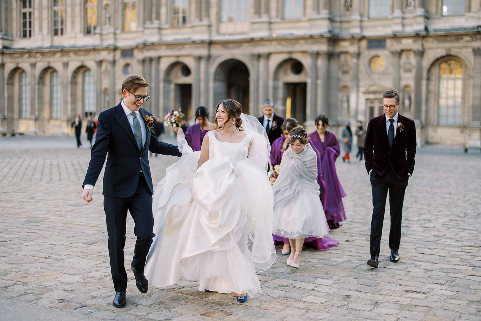 The bride and groom walk across a cobblestone courtyard in front of a grand classical French stone building with arched windows and ornate architectural detailing, likely the Palace of Versailles or a similar historic Parisian monument. The bride wears a voluminous white ball gown with a square neckline, layered skirt, cathedral-length veil, and blue heeled shoes, while the groom wears a navy suit with a grey tie and a floral boutonnière. Behind them walk bridesmaids in deep purple floor-length wrap capes or coats, a young flower girl in a white dress with a grey shawl, and a groomsman in a dark burgundy velvet blazer with black trousers. The group appears to be in motion after a ceremony, laughing and relaxed, captured in a wide candid portrait shot with the historic facade softly out of focus in the background.
