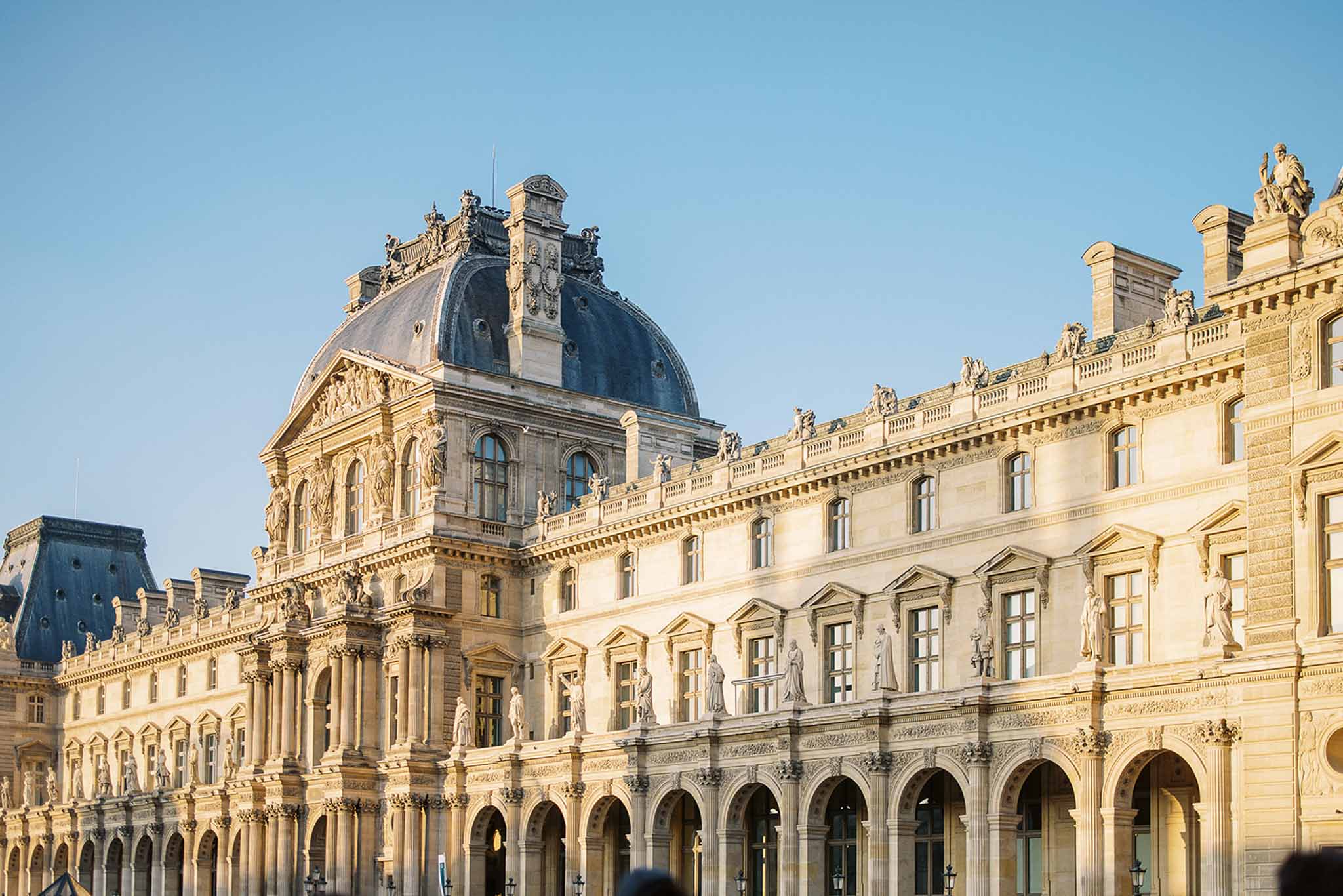 A wide-angle exterior shot of the Louvre Palace courtyard facade in Paris, showing the classic French Renaissance architecture with its cream-colored stone, ornate sculptural detailing, arched ground-floor colonnades, and a dark slate-domed pavilion rising at the wing junction. The building features multiple tiers of tall arched windows, relief sculptures, and full-figure statues positioned between bays across two upper floors. No people, couple, or wedding activity are visible in this image — it functions purely as an architectural location shot. Potential venue feature image.