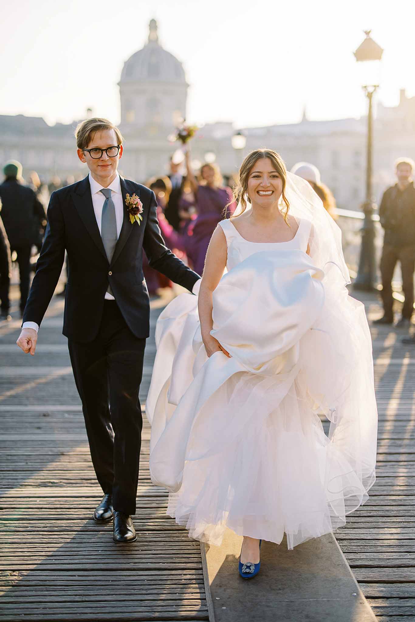 The bride and groom walk together along the Pont des Arts pedestrian bridge in Paris, with the dome of the Institut de France visible in the background. The bride wears a voluminous white ballgown with a square neckline and a long veil, and has paired the look with cobalt blue embellished heels; she is holding up her skirt and smiling broadly at the camera. The groom wears a navy suit with a grey tie and a boutonnière featuring dark burgundy blooms and small berries. A group of guests follows behind them in the warm late-afternoon golden light, with several guests in jewel-toned outfits including fuchsia and deep purple. This is a wide portrait shot capturing the couple mid-stride with the iconic Parisian skyline providing context in the soft-focus background.