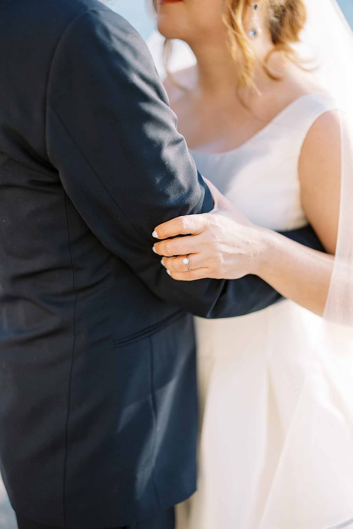 A close-up portrait of a bride and groom embracing during what appears to be a couples portrait session outdoors in bright natural light. The bride, wearing an ivory cap-sleeve gown and a sheer veil, rests her hand on the groom's arm, prominently displaying a round solitaire diamond engagement ring on a delicate band. The groom is dressed in a dark navy suit. The composition is tightly cropped at torso level, with both faces partially visible at the top of the frame, keeping the focus on the couple's hands and the connection between them.
