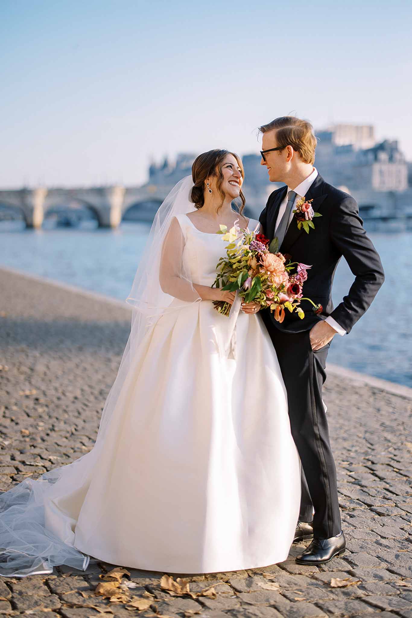 A couple portrait taken outdoors along the Seine riverbank in Paris, with a stone bridge and Haussmann-era buildings visible in the background. The bride wears an ivory satin ballgown with a square neckline, elbow-length veil, and dark drop earrings, holding a loose, garden-style bouquet of coral dahlias, burgundy anemones, mauve blooms, and mixed greenery. The groom wears a dark navy suit with a white dress shirt, grey tie, and a matching boutonniere of burgundy and coral flowers with greenery. The couple stands close together on cobblestones, smiling at each other, shot as a full-length portrait with warm golden-hour side lighting.
