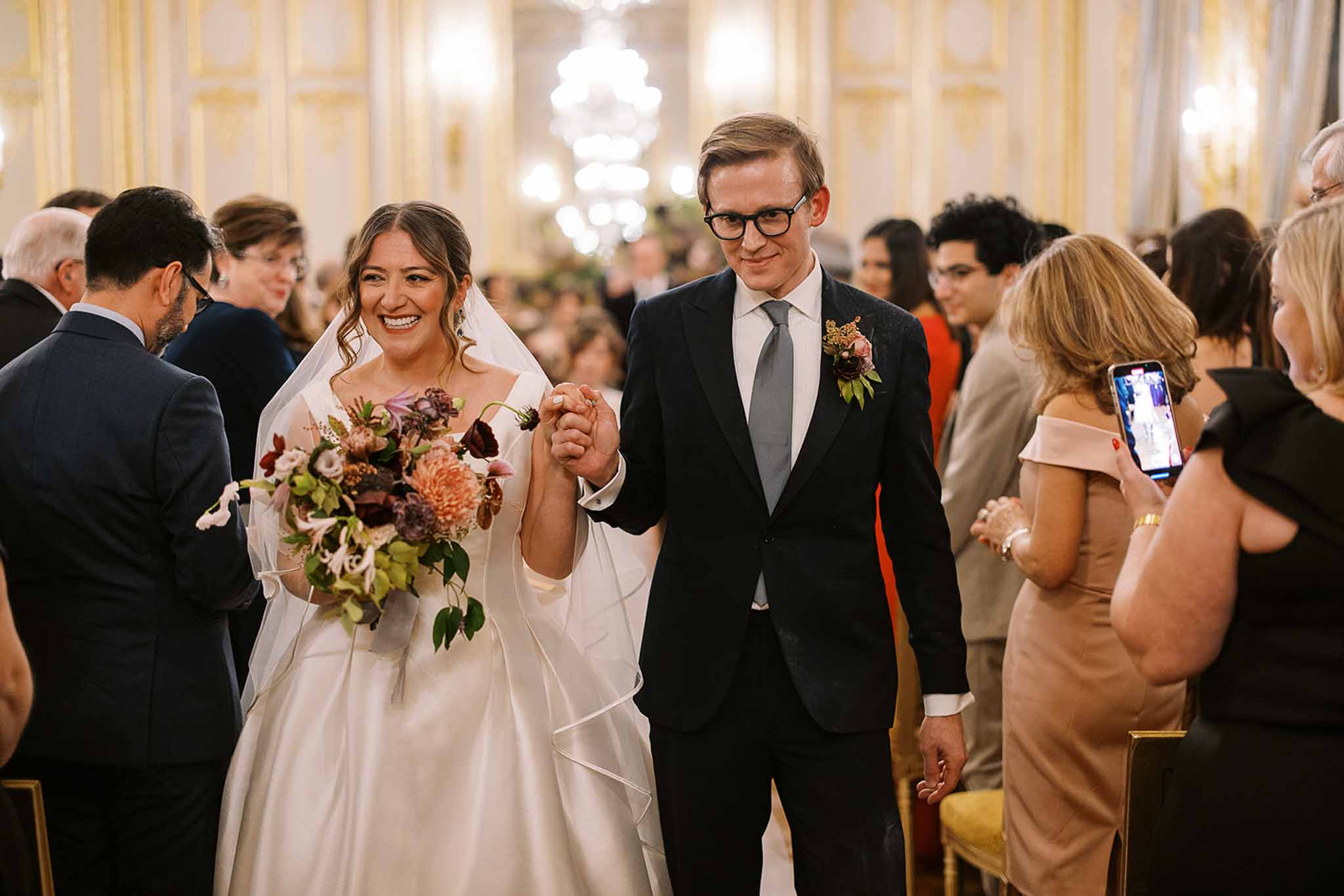 The bride and groom walk back down the aisle together following their indoor ceremony, holding hands and smiling broadly. The setting is a grand ballroom-style venue with ornate gold-and-white paneled walls, crystal chandeliers, and gilded chairs lining the aisle, suggesting a classic Parisian or chateau interior. The bride wears an ivory satin ball gown with a long veil and carries a lush, oversized bouquet featuring deep burgundy dahlias, blush chrysanthemums, green cymbidium orchids, dark purple anemones, and trailing foliage in an autumn-toned palette; the groom wears a navy suit with a sage green tie and a boutonniere echoing the bouquet's tones with blush roses and berries. Approximately 30 or more guests are visible on both sides of the aisle, several turning to watch or photograph the couple, captured in a mid-shot portrait with soft warm ambient lighting from the chandeliers.