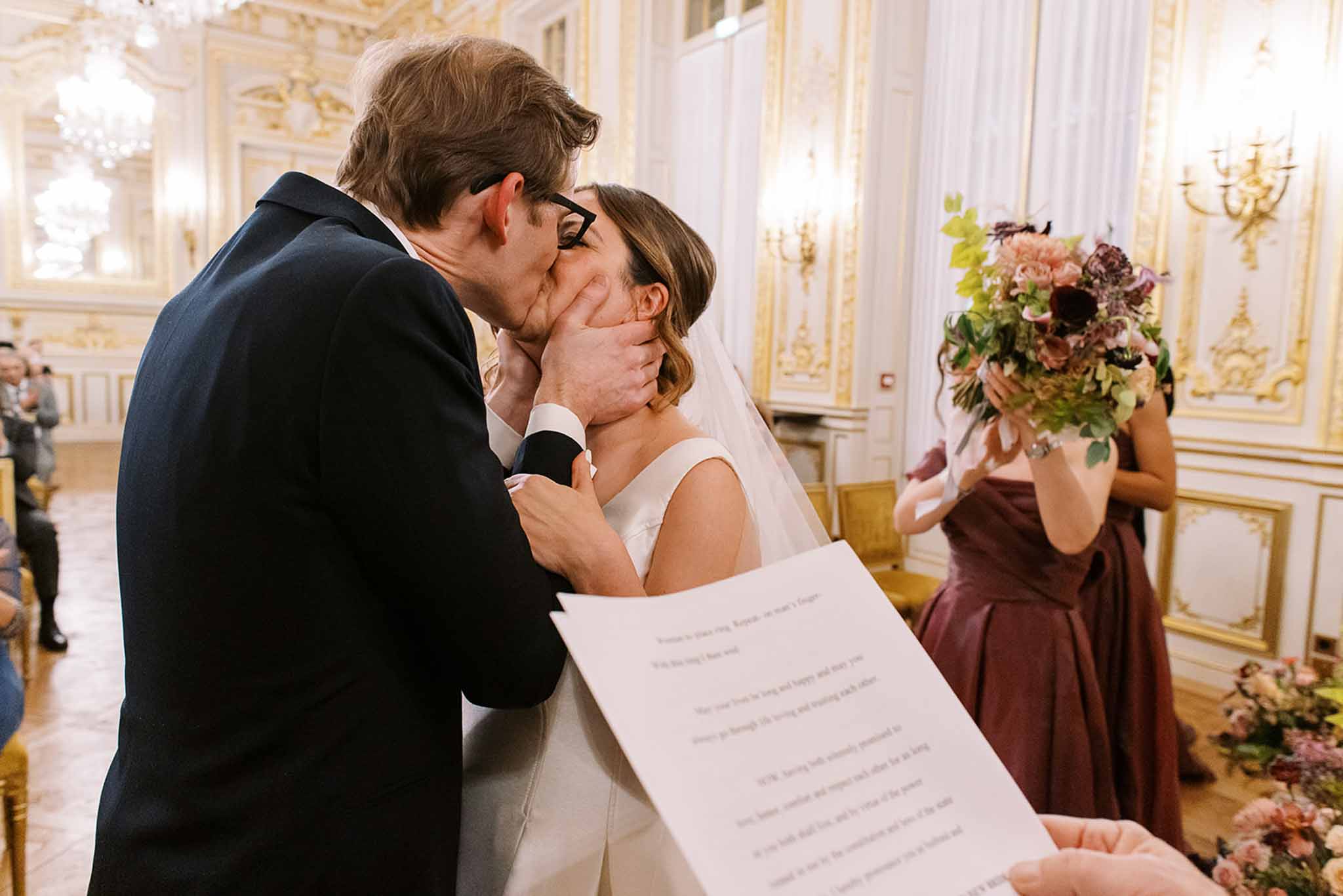 The couple shares their first kiss during an indoor civil or religious ceremony, taking place in a ornate ballroom-style venue with gold-trimmed white paneling, crystal chandeliers, and wall sconces — consistent with a Parisian hôtel particulier or mairie. The groom, wearing a navy suit and black-framed glasses, cups the bride's face with both hands as they kiss; the bride wears a sleeveless ivory gown with a veil. In the foreground, partially out of focus, a hand holds a printed ceremony reading. To the right, a bridesmaid in a deep burgundy/mauve ball-style dress holds a bouquet of blush pink, dusty mauve, and deep plum florals with greenery, while additional floral arrangements in similar tones are visible at the ceremony's edge. Seated guests are visible in the background on the left. The shot is taken at mid-range from a slightly low angle, capturing the emotional moment with the ornate venue decor visible throughout the frame.