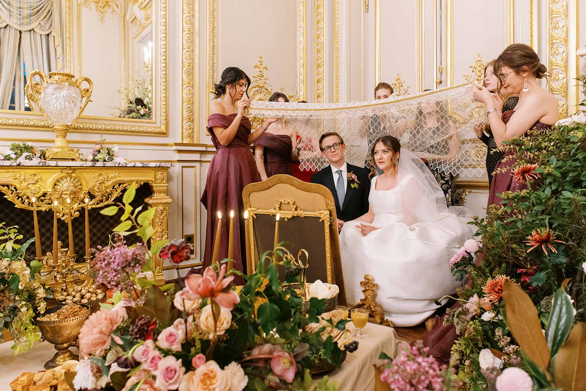 A Jewish wedding ceremony is taking place indoors in an ornate French-style ballroom or salon with cream and gold-gilded paneling, decorative moldings, and a large gold-framed fireplace. The bride, seated in a gold carved chair, wears a white ball gown with a lace-trimmed veil, while the groom sits beside her in a dark suit with a light blue tie and a floral boutonniere; several bridesmaids in off-the-shoulder burgundy/deep wine gowns hold a white lace chuppah canopy overhead. In the foreground, the ceremony table is densely arranged with blush, coral, peach, and pink garden roses, dahlias, and amaryllis blooms with abundant greenery, gold candelabras, wine glasses, and ritual objects including what appear to be wedding rings on a decorative holder. The setting has a classic, formal aesthetic with rich floral arrangements in warm autumnal tones throughout the room. Wide-angle shot capturing both the foreground ceremony table details and the seated couple with attendants behind.