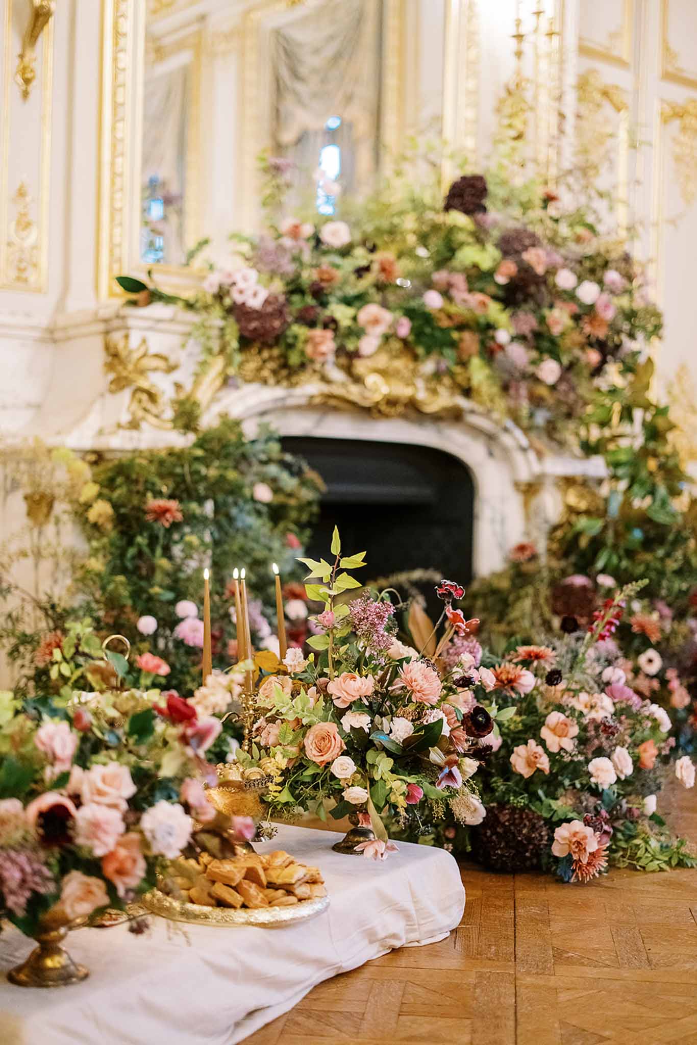A reception tablescape and floral installation photographed indoors in a grand ballroom or salon featuring ornate white and gold rococo paneling and a large carved marble fireplace. The focal point is a sweeping floral arrangement that cascades from the mantelpiece down to the floor, composed of blush and peach garden roses, burgundy dahlias, mauve and dusty pink blooms, deep plum anemones, rust-toned flowers, and abundant mixed greenery in an abundant, garden-gathered style. In the foreground, a white linen-draped table holds a gold footed tray of pastries or canapes, gold candelabras with lit taper candles, and additional floral arrangements continuing the same warm, autumnal palette. The herringbone parquet wood floor is visible beneath the arrangements. The overall decor style is maximalist and romantic with a rich, jewel-toned floral palette against a classical French interior. Medium wide shot capturing both the table details and the full fireplace backdrop. Potential venue feature image.