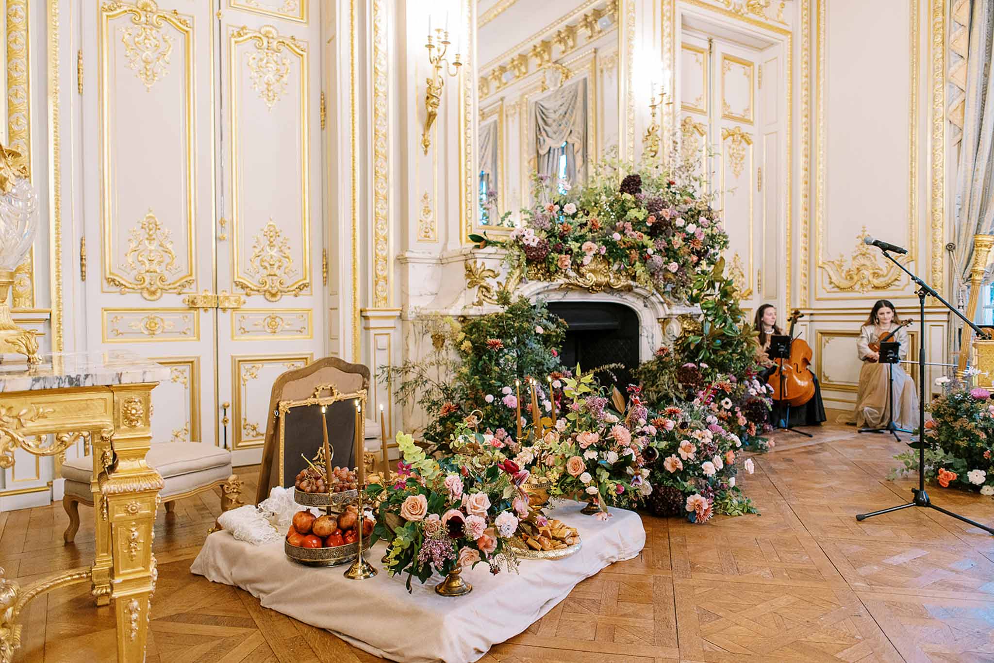 A wedding reception setup photographed indoors in a grand French ballroom or salon with white and gold boiserie paneling, a white marble fireplace, and herringbone parquet flooring. The focal point is a large floor-level floral installation cascading from the top of the fireplace mantel down to the floor, composed of blush pink roses, coral garden roses, burgundy dahlias, soft lilac blooms, greenery, and trailing foliage in an abundant, garden-style arrangement. In the foreground, a low table draped in a white cloth holds brass candlesticks with tapered gold candles, a tiered stand of grapes, a bowl of pomegranates, and a plate of pastries, suggesting a sofreh aghd or Persian-inspired ceremony table. To the right, two female musicians — one with a cello and one with a violin — are seated and performing, both dressed in neutral-toned gowns. The wide shot captures the full room setup with gilded Louis XVI-style furniture visible on the left. Potential venue feature image.