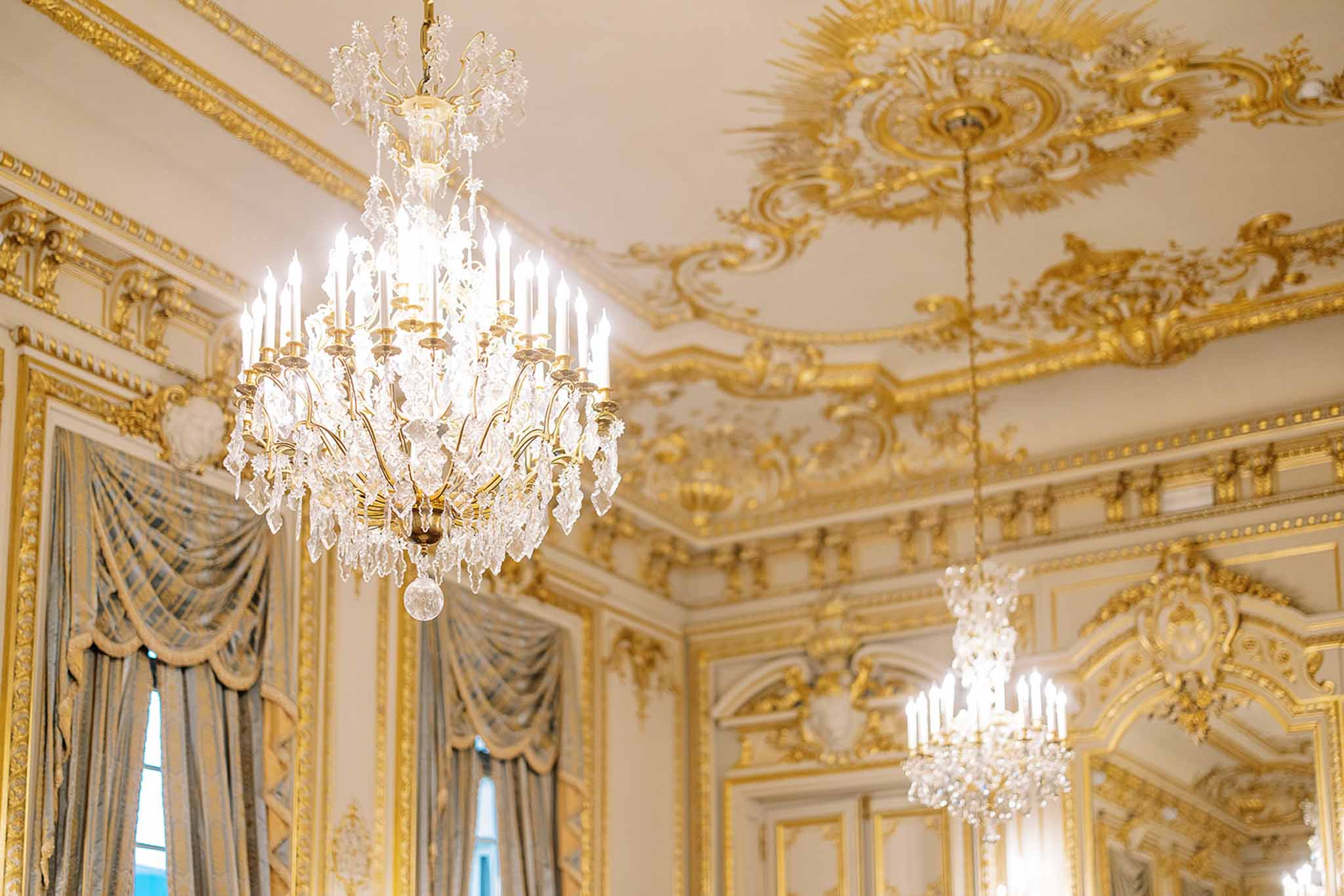 A detail/close-up interior shot of a formal ballroom or reception hall featuring two large crystal and gold chandelier candelabra-style light fixtures hanging from a white ceiling with heavily gilded Rococo-style plasterwork, including sunburst and scrollwork ceiling medallions. The walls are paneled in white with ornate gold molding throughout, and draped sage-green silk curtains with gold-trimmed framing are visible along the left side. No people are present in the image. The overall decor palette is white, gold, and sage green, consistent with a classic French palatial interior. Potential venue feature image.