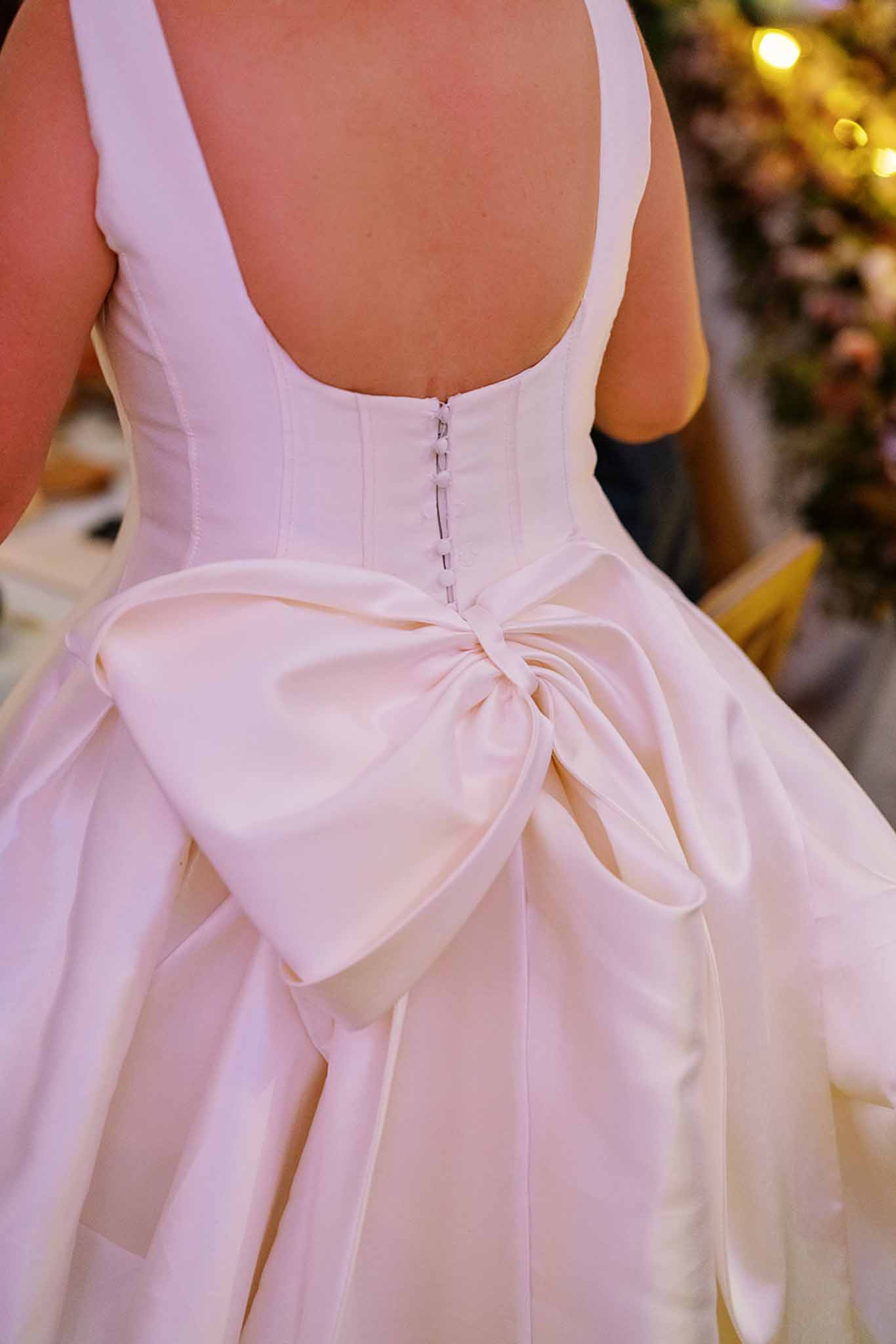 Close-up detail shot of the back of a bride's ivory satin ball gown, featuring a structured boned corset bodice with a low open back, hook-and-eye closure, and a large oversized satin bow at the waist. The full skirt falls in voluminous pleats below the bow. The background is blurred but suggests an indoor reception setting with warm golden lighting and greenery, with guests visible out of focus.