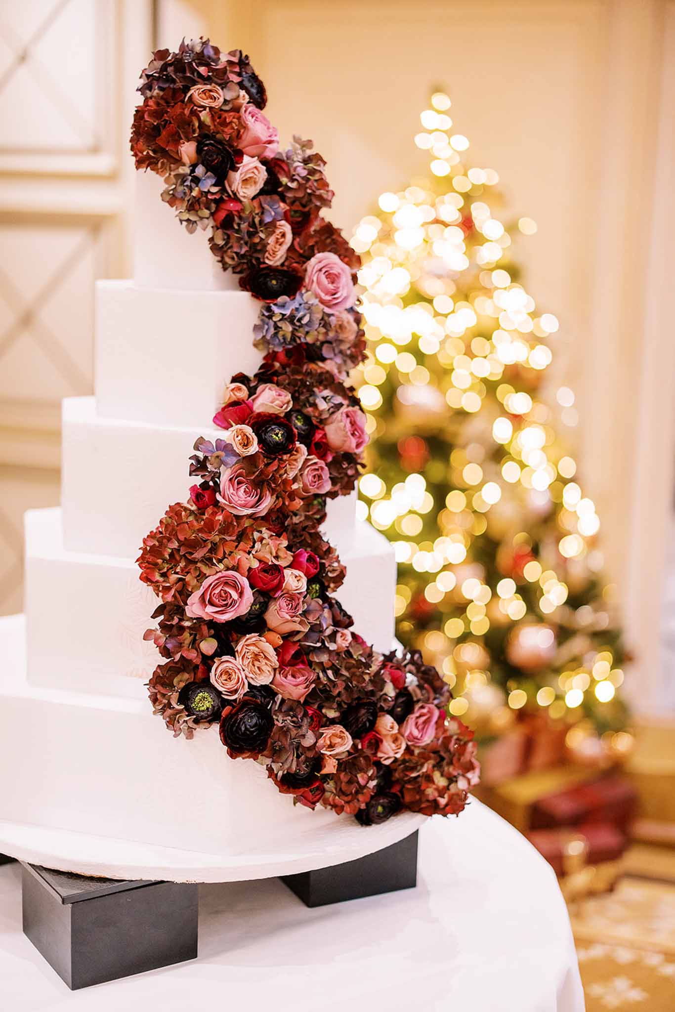 Close-up detail shot of a five-tier white fondant wedding cake displayed on a white-draped table with black risers, set indoors in what appears to be a ballroom or reception hall. A dramatic cascading floral arrangement runs diagonally across the cake from base to top, composed of deep burgundy dried hydrangeas, blush and mauve garden roses, dark navy-black ranunculus, and touches of dusty blue hydrangeas. In the soft-focus background, a fully lit Christmas tree with warm golden bokeh lights suggests a winter or holiday-season wedding. The overall decor palette is deep jewel-toned and rich, consistent with a formal winter wedding aesthetic.
