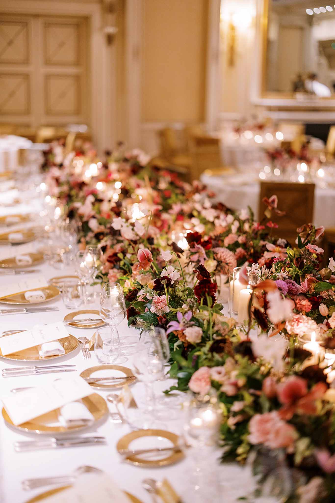 A detail shot of a wedding reception tablescape inside a formal ballroom with warm cream-toned paneled walls and gold wall sconces. The long banquet table is dressed in a white linen tablecloth and set with gold charger plates, folded white napkins, silver flatware, and multiple crystal wine glasses per place setting. A dense, low floral runner stretches the full length of the table, composed of deep burgundy ranunculus and anemones, blush and dusty rose garden roses, pink carnations, mauve chrysanthemums, and small purple orchids interspersed with greenery. Pillar candles in glass vessels are tucked throughout the floral runner, providing warm candlelight. Additional round tables with matching gold charger settings and floral centerpieces are visible in the soft background. The overall decor palette is deep jewel-toned florals against white and gold, consistent with a classic, formal French ballroom reception styling.