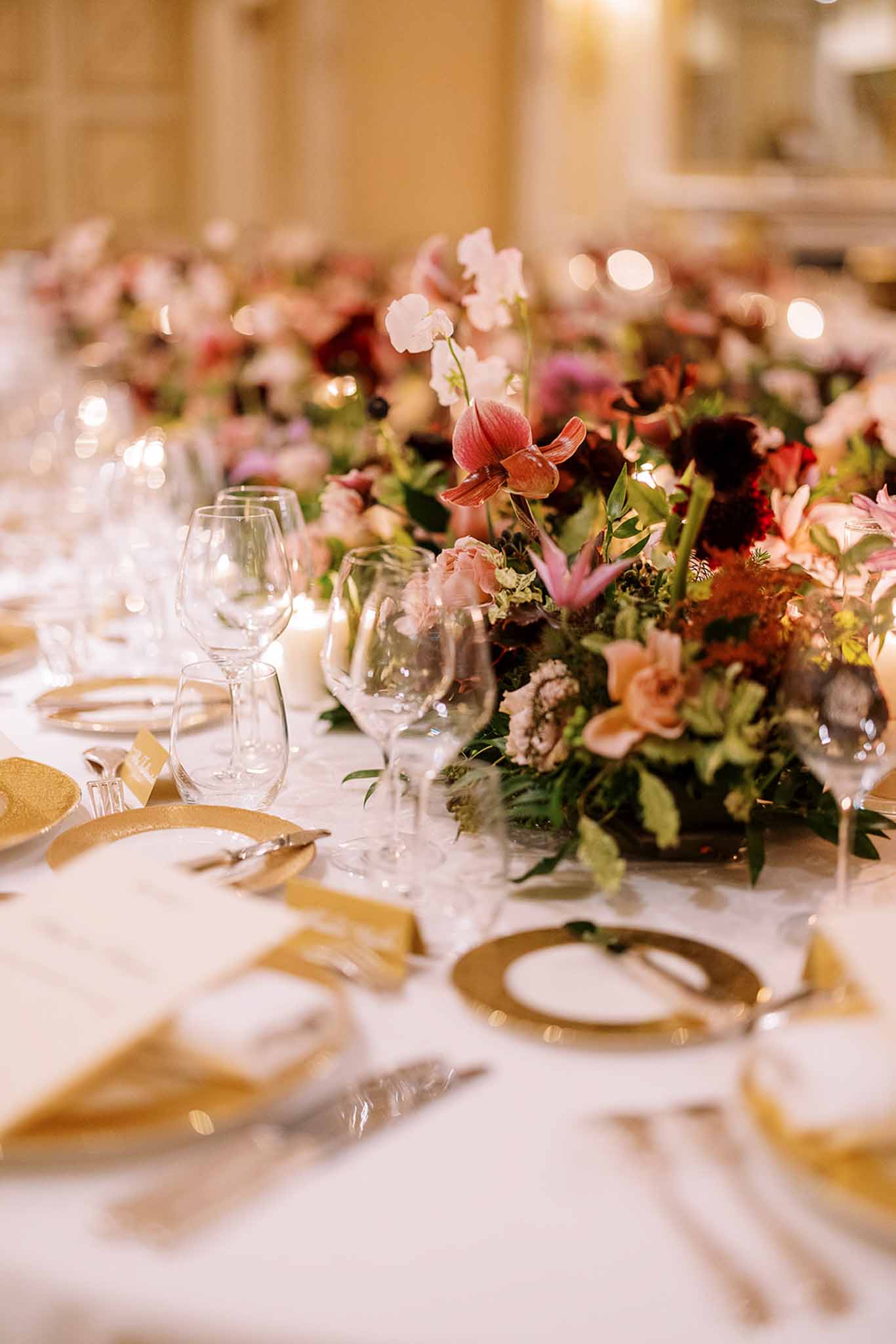 Close-up detail shot of a wedding reception table setting inside a warmly lit ballroom or formal indoor venue. The long table is dressed with a white linen and set with gold charger plates, silver cutlery, small gold place cards, and multiple clear wine and water glasses per setting. A low, lush floral centerpiece runs along the table featuring blush pink sweet peas, coral anthurium, deep burgundy dahlias, blush garden roses, pink lilies, and mixed greenery in a rich, jewel-toned garden-style arrangement. The decor palette combines gold, white, and deep berry tones, reflecting a classic formal styling theme. Additional centerpieces are visible in soft focus extending down the length of the table, with warm ambient lighting visible in the background.