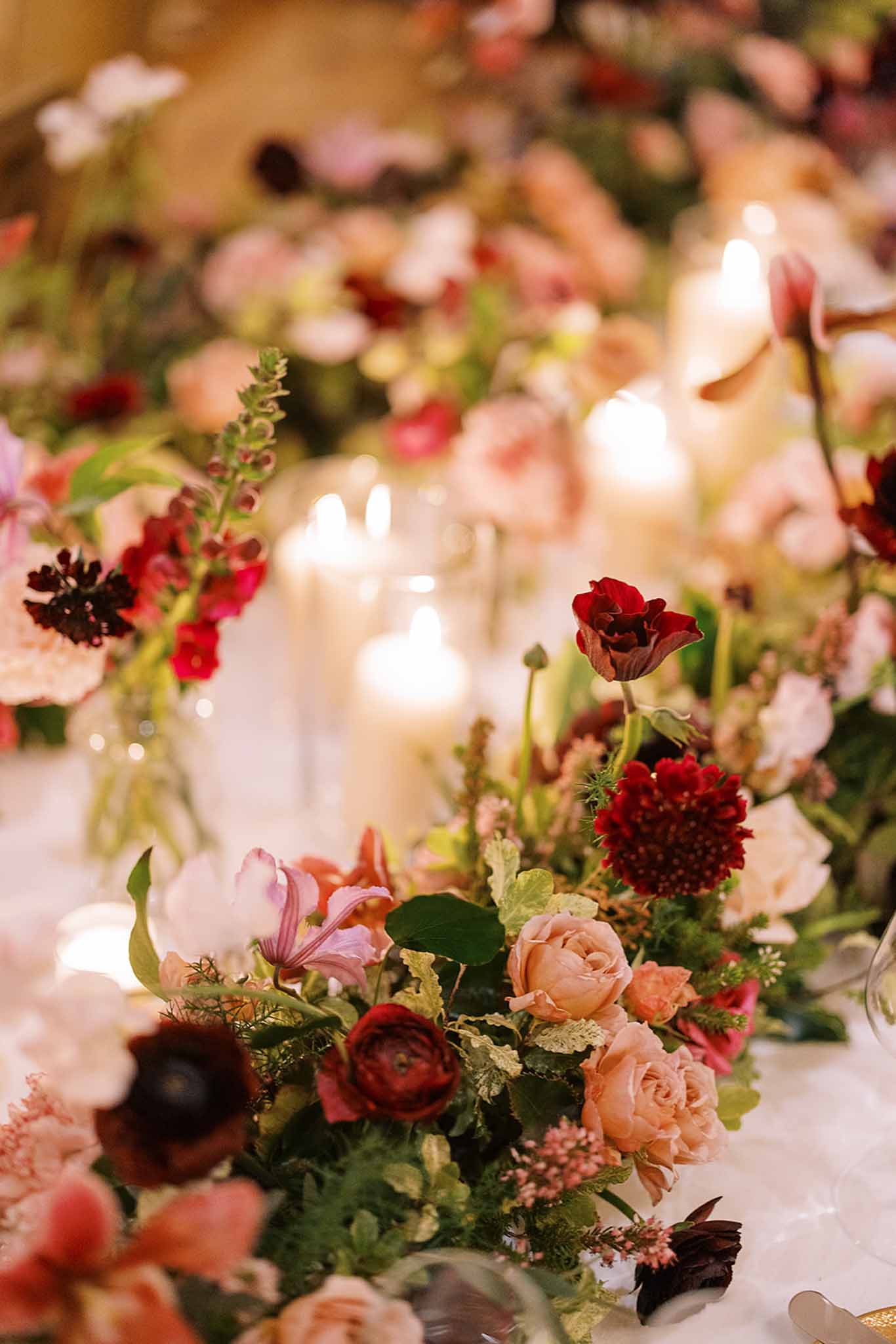 Close-up detail shot of a wedding reception table runner featuring a lush, garden-style floral arrangement with deep burgundy ranunculus, dark red poppies, blush garden roses, dark crimson scabiosa, pink cosmos, and mixed greenery including ferns and textured foliage. The color palette ranges from blush and peach to deep wine and burgundy tones, with scattered pink waxflower adding softness throughout. Multiple lit taper candles in glass holders run along the center of the table, casting a warm, intimate glow across the arrangement. The table is covered in a white linen, and the composition uses a shallow depth of field, keeping the foreground flowers sharp while the background candles and blooms blur softly.