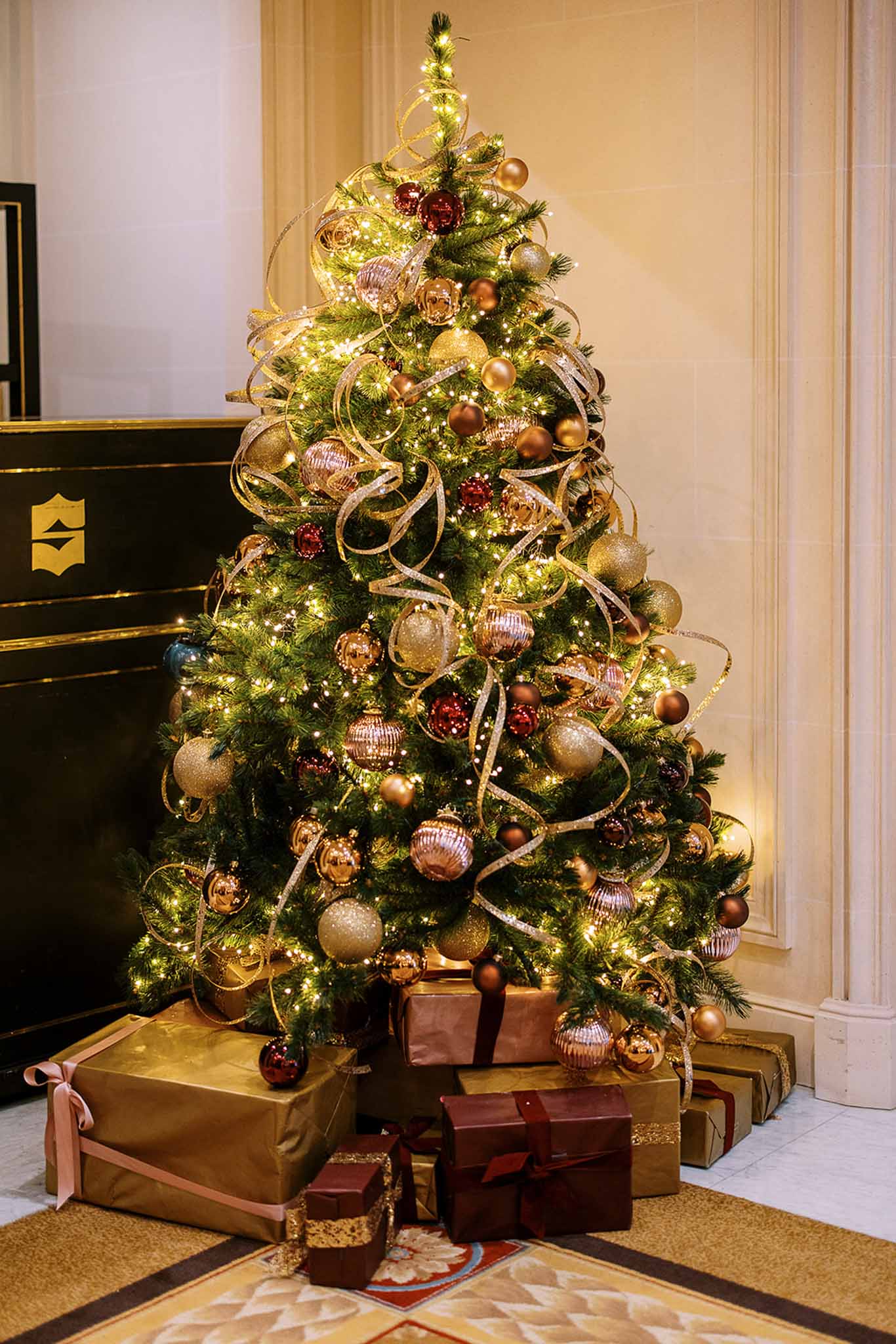 This image does not depict a wedding photograph. It shows a decorated Christmas tree in an indoor setting, likely a hotel lobby or formal venue interior, with cream-colored paneled walls and a dark reception desk with a gold emblem visible to the left. The tree is decorated with warm white fairy lights, gold glitter ribbon, and a mix of gold, rose gold, burgundy, and champagne ornaments in varying sizes and finishes. Wrapped gifts in gold, burgundy, and rose packaging are arranged beneath the tree on a patterned area rug. The overall decor palette is gold and burgundy with warm ambient lighting. Wide shot, slightly low angle.