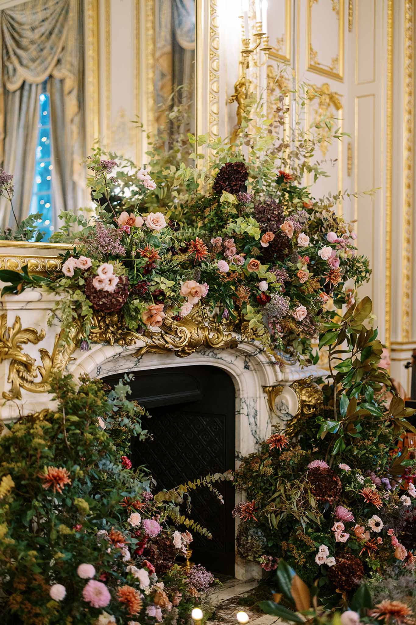 A close-up detail shot of an oversized floral installation completely engulfing a white marble and gilt fireplace inside a grand French interior, likely a historic chateau or palace-style reception venue. The arrangement features blush and antique pink garden roses, deep burgundy and mocha hydrangeas, burnt orange and rust dahlias and chrysanthemums, mauve lilac clusters, and abundant trailing greenery including eucalyptus, ivy, and magnolia leaves. The palette is rich and autumnal with warm terracotta, deep plum, blush, and green tones, styled in a lush, overflowing botanical aesthetic. The room behind features cream and gold boiserie wall paneling, a gilded candelabra, and blue-grey silk drape curtains with fairy lights visible through the window, indicating an evening reception setting.
