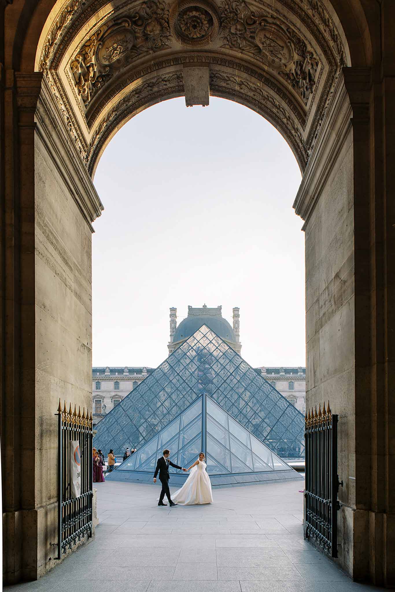 A couple's wedding portrait shoot at the Louvre Museum in Paris, photographed from a distance through one of the museum's ornately carved stone archways. The bride wears a full-length ivory ballgown with a trailing skirt, and the groom is dressed in a dark suit; they are holding hands and walking across the courtyard plaza in front of the iconic glass pyramid. The composition frames the couple as small figures centered within the arch, with the Louvre pyramid and the classical Haussmann-style museum facade visible in the background, along with a small group of tourists to the left. The wide shot uses the architectural framing of the arch to create strong perspective depth, with warm late-afternoon light casting a golden tone on the stone surroundings. Potential venue feature image.