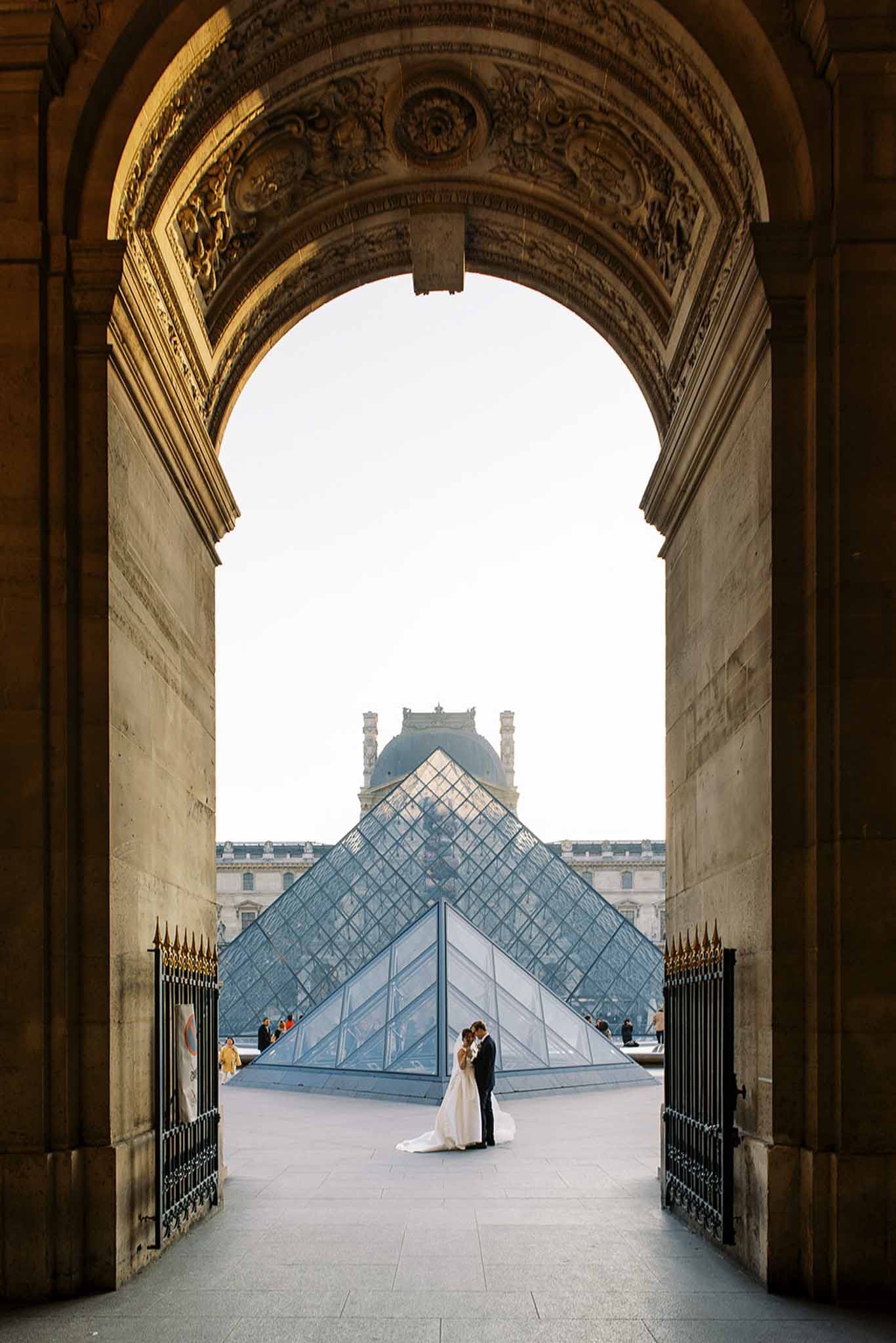 A couple portrait taken outdoors at the Louvre Museum in Paris, framed through a large ornate stone archway with detailed carved relief work. The bride wears a white ball gown with a cathedral-length train and the groom is dressed in a dark suit; the two are standing close together, facing each other, in front of the iconic glass pyramid. The composition is a wide shot that uses the archway as a natural frame, placing the couple small in the middle distance with the pyramid and the classical Louvre palace facade visible behind them. A small number of other visitors are visible in the background near the pyramid. Potential venue feature image.