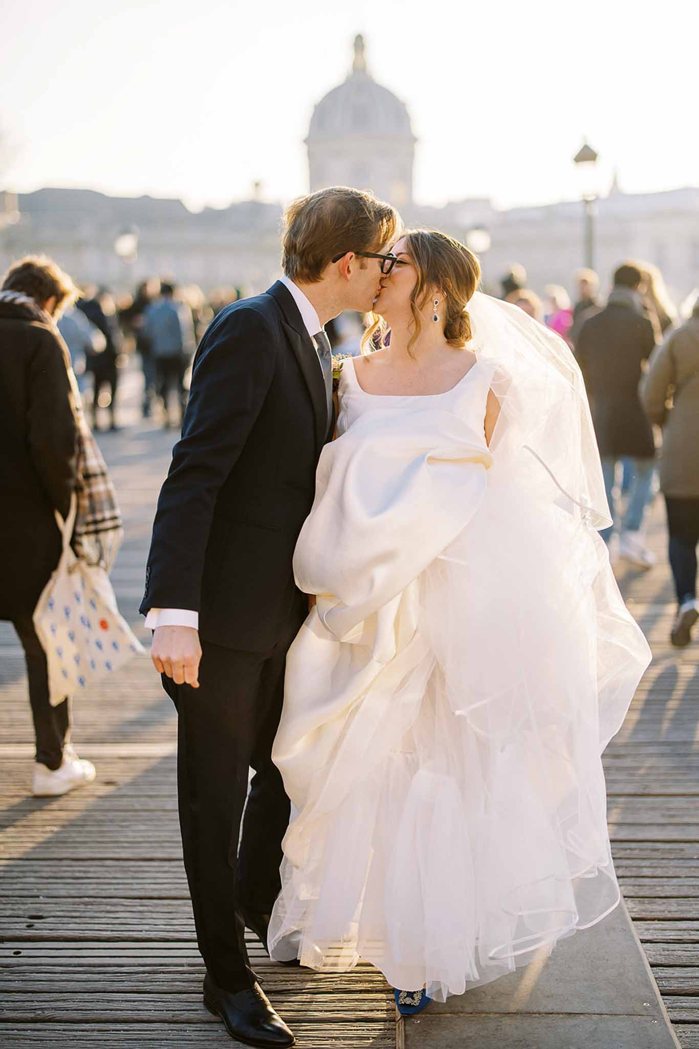 A couple portrait taken outdoors on the Pont des Arts bridge in Paris, with the Institut de France dome visible in the soft-focus background. The bride and groom are kissing in the middle of the busy pedestrian bridge, surrounded by passersby. The groom wears a dark navy suit with a tie and black-framed glasses, while the bride wears an ivory ball gown with a square neckline, a voluminous layered organza skirt, a white wrap or cape, a long veil, sapphire drop earrings, and cobalt blue embellished heels visible beneath the dress. The shot is a medium full-length portrait taken at golden hour, with warm backlight creating a halo effect around the couple.
