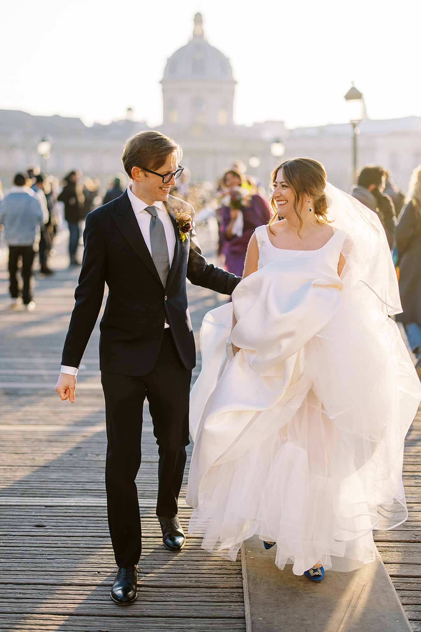 A couple portrait taken outdoors on the Pont des Arts pedestrian bridge in Paris, with the dome of the Institut de France visible in the soft-focus background. The groom wears a dark navy suit with a grey tie and black-rimmed glasses, while the bride wears a voluminous white ballgown with a square neckline, a long veil, blue embellished heels, and blue drop earrings. The groom holds the bride's train as they walk and laugh together, with a crowd of passersby visible behind them in warm golden-hour light. The shot is a full-length walking portrait with a shallow depth of field emphasizing the couple against the iconic Parisian backdrop.