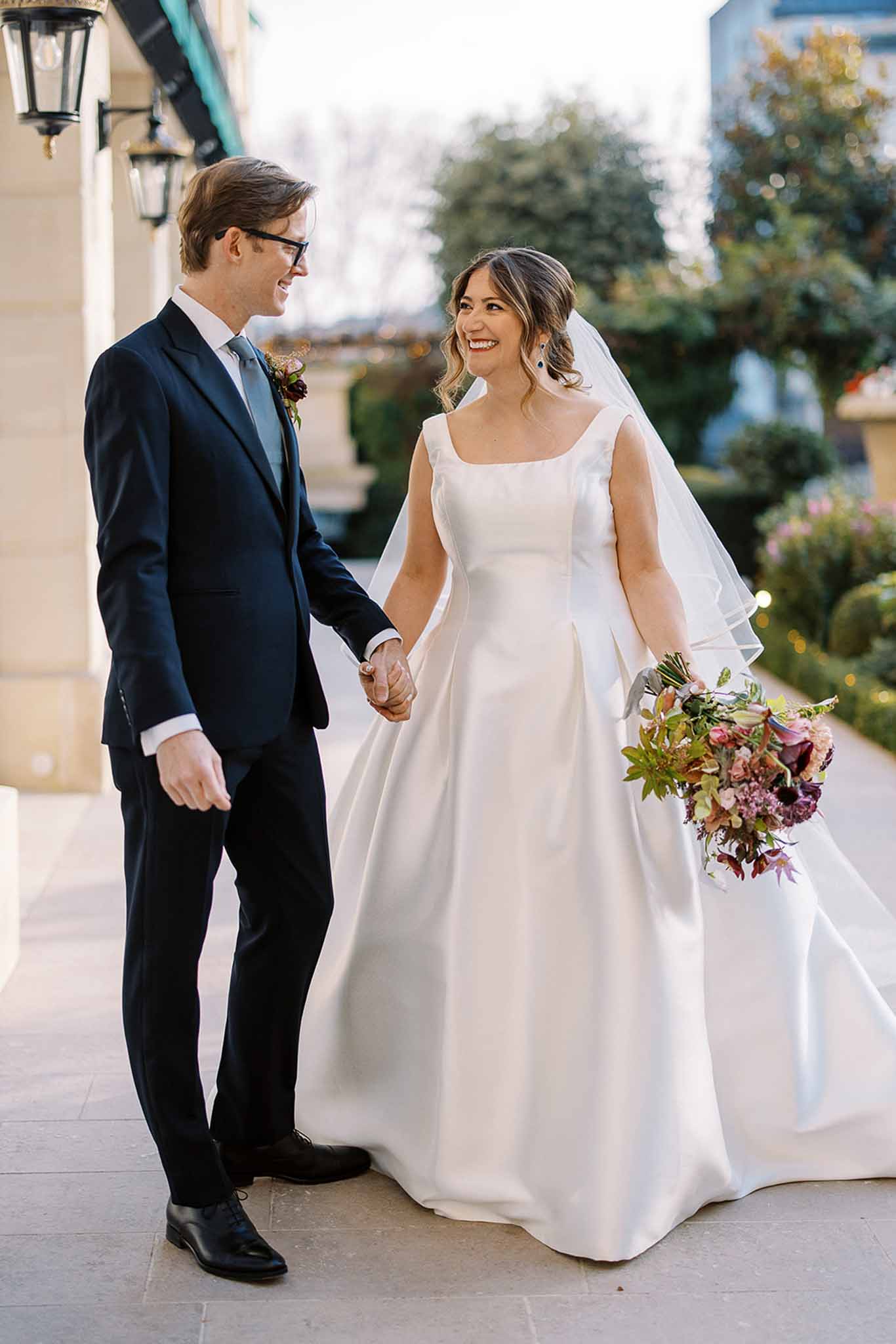 A couple portrait taken outdoors on a paved terrace beside a classical building with wall-mounted lanterns visible in the background. The groom wears a navy suit with a grey-blue tie, black-framed glasses, and a small burgundy boutonniere, while the bride wears a white satin ball gown with a square neckline and a long veil with a rolled edge. The bride carries a loosely arranged bouquet in deep burgundy, mauve, rust, and blush tones with mixed foliage. The two are holding hands and looking at each other, both smiling. The composition is a full-length couple portrait with a shallow depth of field that blurs the garden setting behind them. The overall styling is clean and modern with rich jewel-toned florals as an accent.