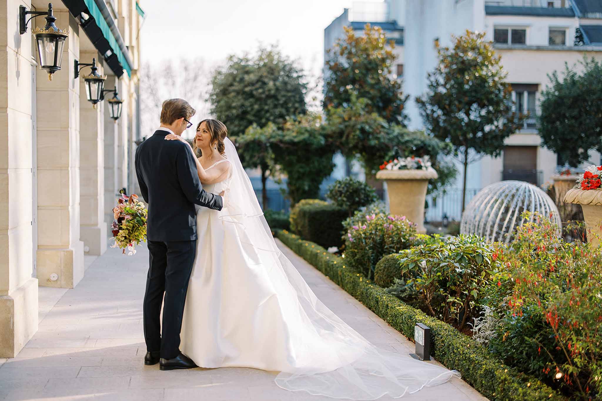 A couple poses for portraits along a terrace walkway beside a Haussmann-style stone building with black iron wall lanterns and a teal awning visible on the left. The bride wears an ivory off-the-shoulder ballgown with a structured bow at the back and a long cathedral-length veil with a trailing hem; the groom wears a dark navy suit with glasses. She faces the camera with her hand on his shoulder while he faces away. The bride holds a richly colored bouquet featuring deep burgundy and red blooms, dark foliage, and trailing greenery. The formal garden beside them includes neatly clipped boxwood hedges, a stone urn planter, and a decorative wire globe sculpture. This is a wide portrait shot with a classic, formal Parisian style.