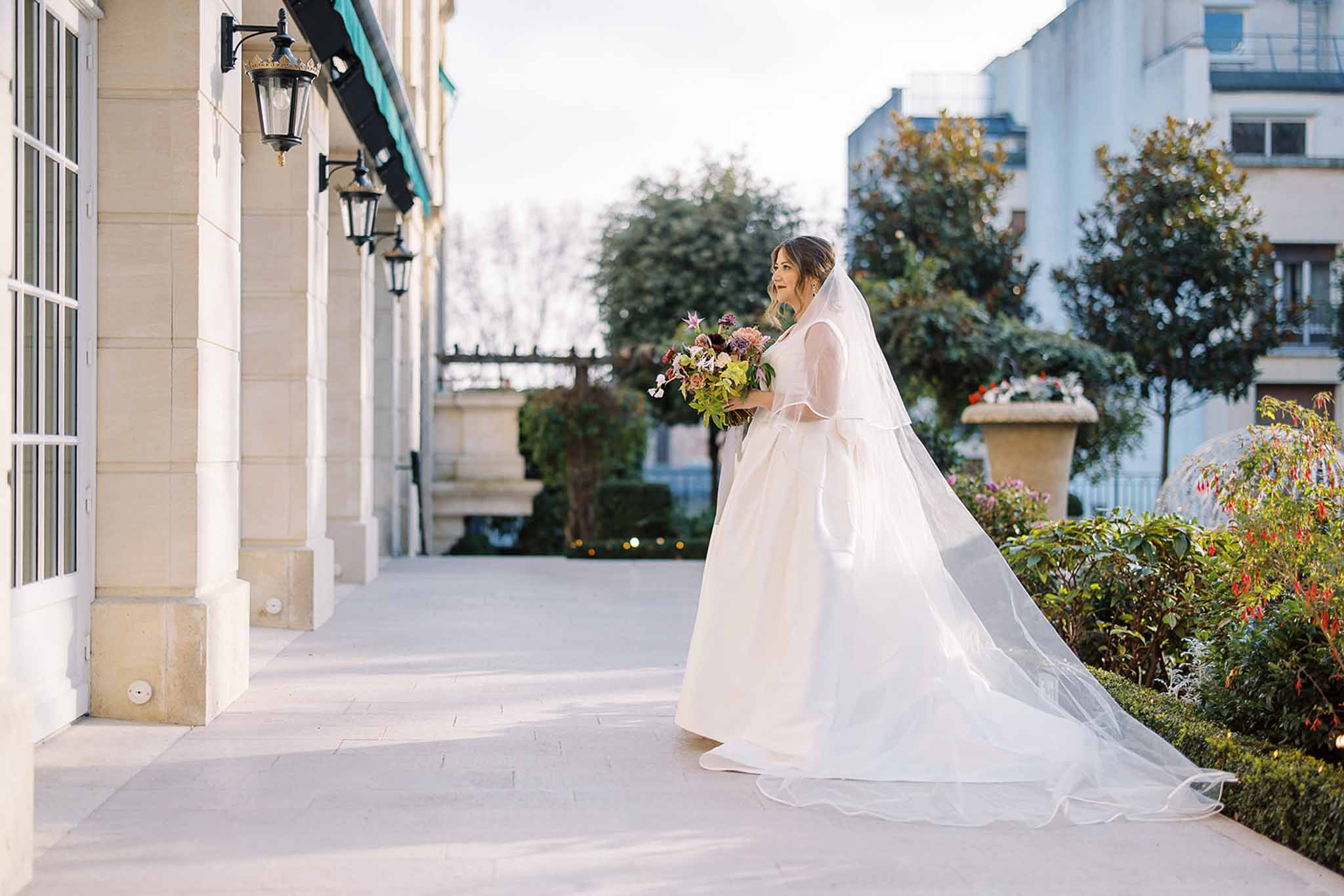 A bridal portrait taken outdoors on a stone terrace alongside a classic Haussmann-style building facade featuring tall windows and black iron lanterns with a teal awning visible above. The bride stands in a full-length ivory ballgown with sheer long sleeves and a cathedral-length two-tier veil that trails along the pavement behind her. She holds a loose, garden-style bouquet featuring deep burgundy, purple, and blush blooms with abundant greenery. The composition is a full-length portrait shot from a slight distance, capturing the full sweep of the veil and gown train against the pale stone terrace.