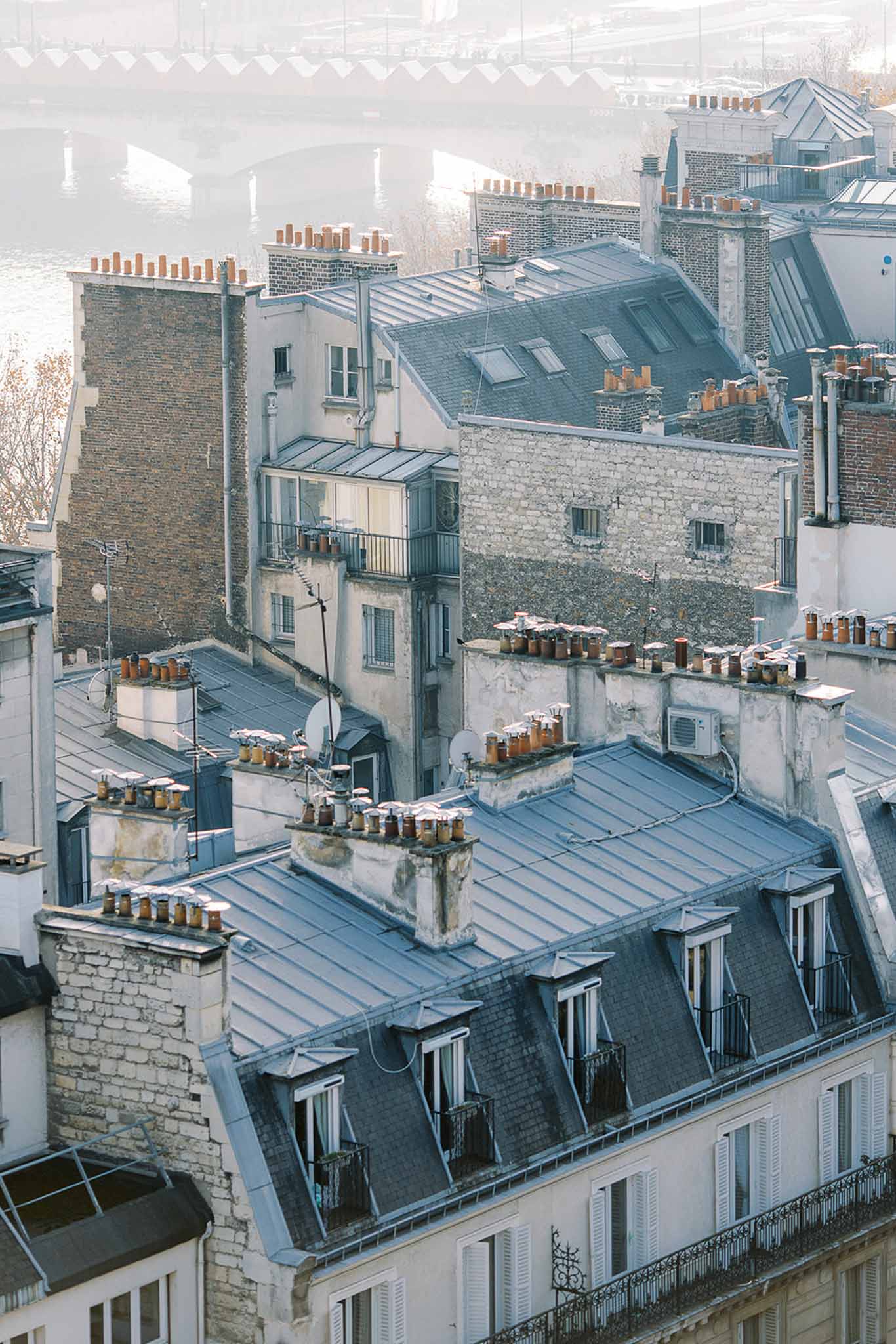 This image contains no wedding-related content. It is an aerial/elevated wide shot of Parisian rooftops featuring classic zinc-grey mansard roofs, terracotta chimney pots, dormer windows, and Haussmann-style building facades in cream and stone. A river — likely the Seine — and a bridge are visible in the misty background. There are no people, couples, wedding attire, florals, or event decor present in the frame.