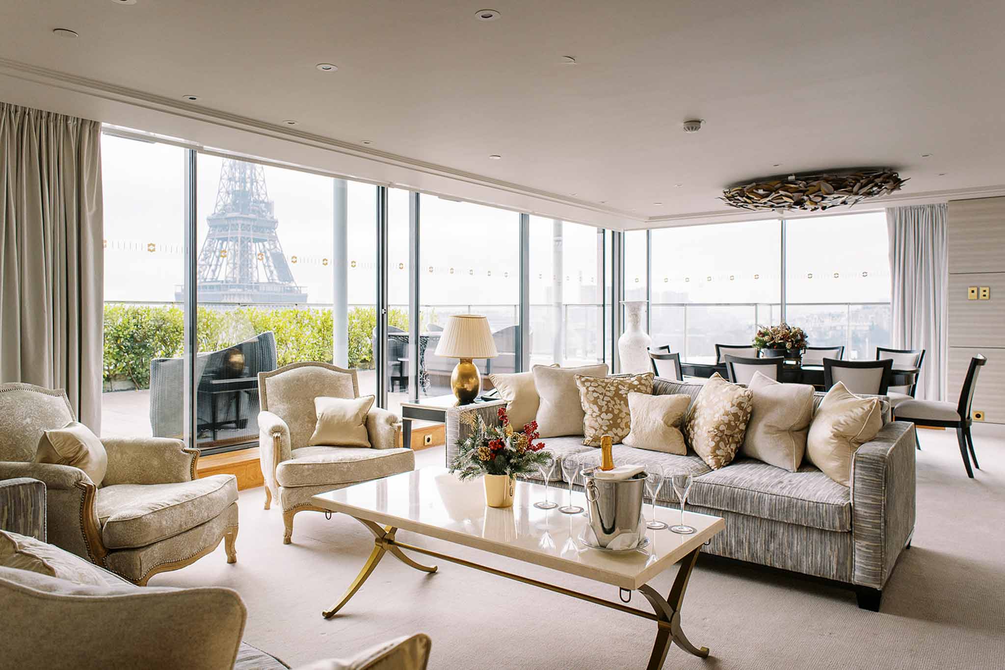 Interior wide shot of a high-end Paris hotel suite or penthouse living room with direct views of the Eiffel Tower through floor-to-ceiling glass windows. The space features a neutral palette of champagne, ivory, and soft grey upholstered furniture including a large sofa, armchairs with carved gilt legs, and a marble-topped coffee table with gold X-frame legs. A champagne bottle in an ice bucket and several champagne flutes are arranged on the coffee table alongside a small holiday floral arrangement with red berries and greenery. A dining area with dark wood chairs and a floral centerpiece is visible in the background. The decor style is classic Parisian with gold accents, a sculptural ceiling fixture, and ivory drape curtains. No people are present. Potential venue feature image.