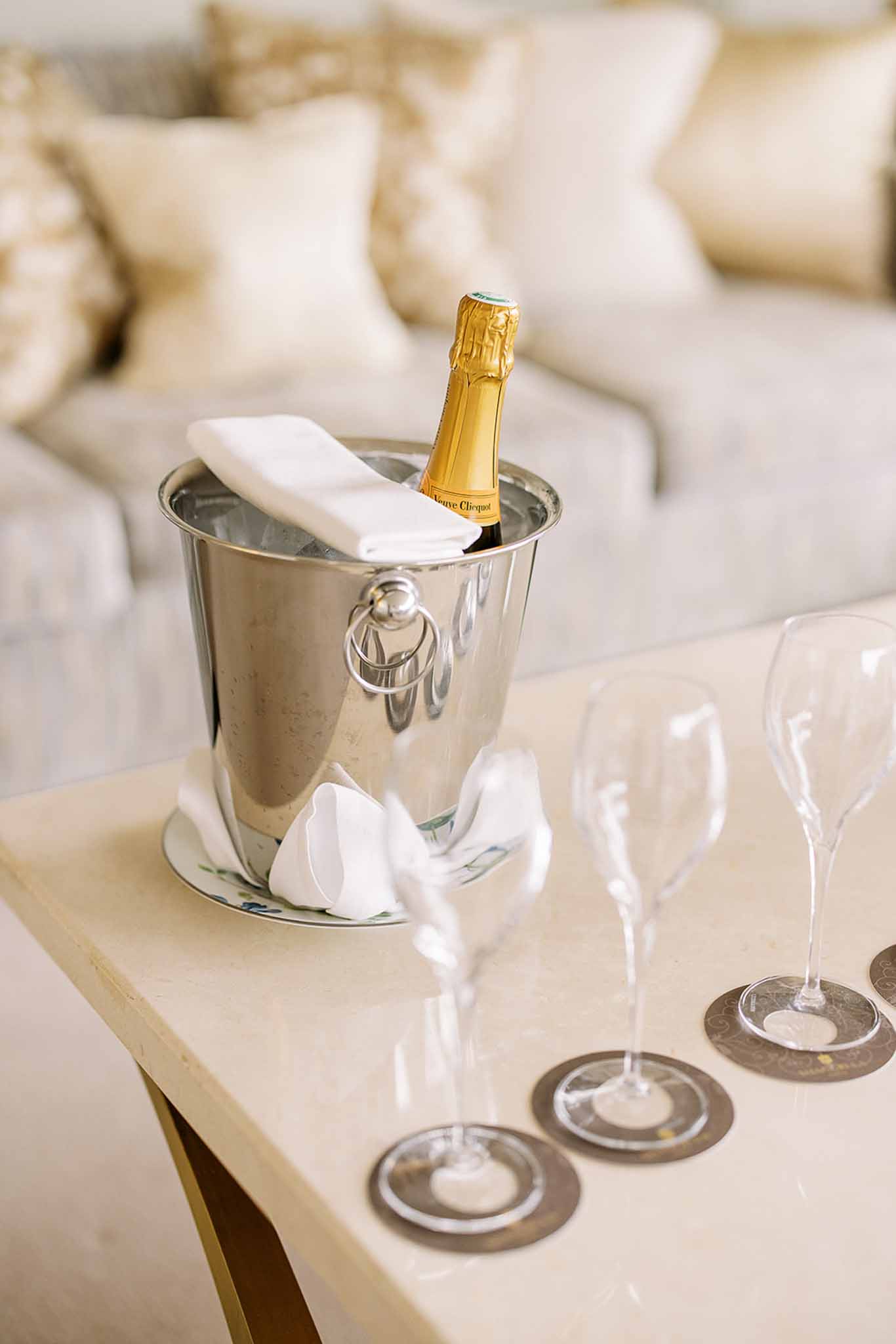A close-up detail shot of a bottle of Veuve Clicquot champagne chilling in a polished silver ice bucket, accompanied by a folded white linen napkin, placed on a cream marble side table. Three tall crystal champagne flutes rest on silver circular coasters beside the bucket. The background shows a cream upholstered sofa with gold and ivory decorative cushions, suggesting an indoor lounge or bridal suite setting with a neutral, ivory-toned decor palette.