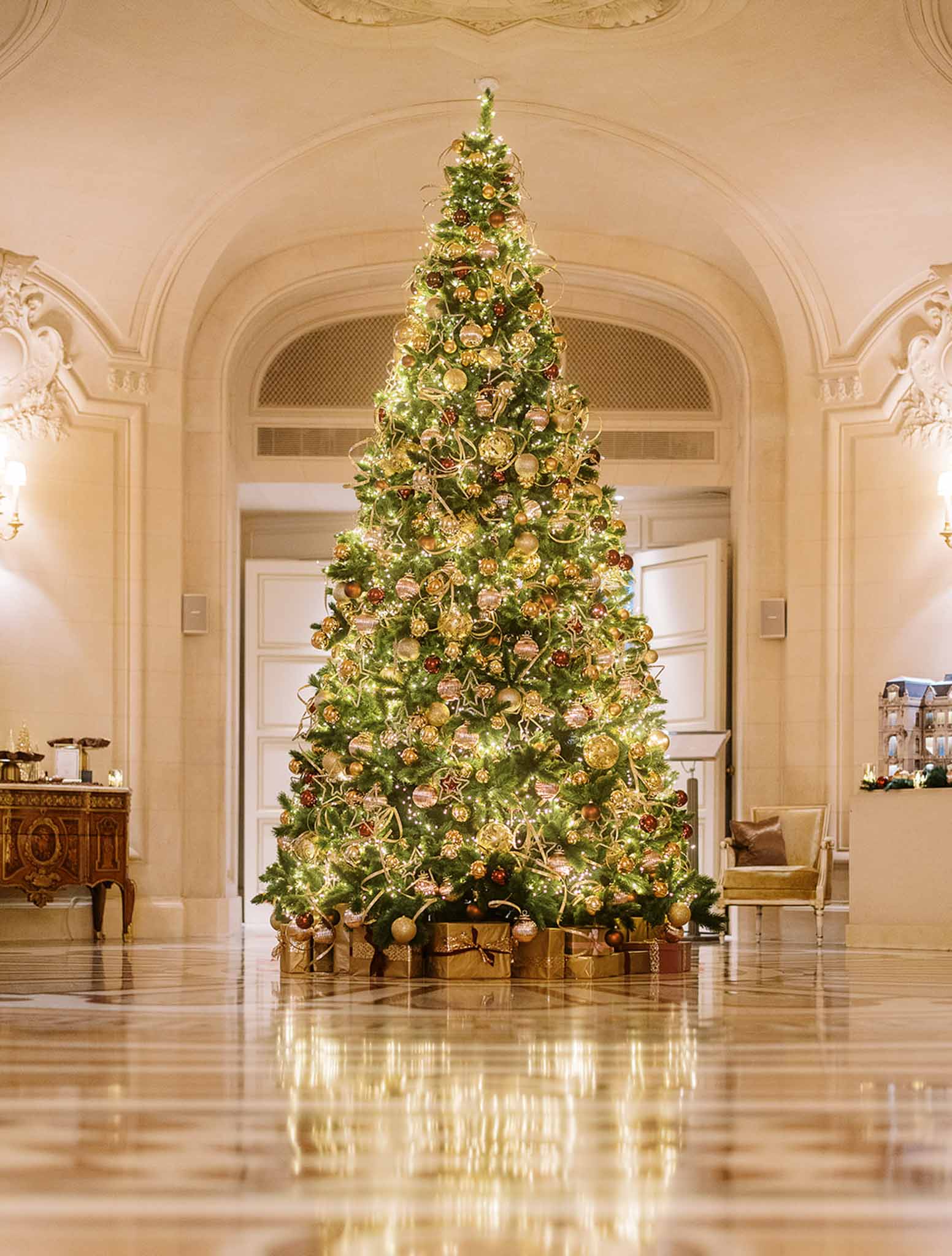 This image does not depict a wedding photograph but rather shows the grand interior foyer of a formal venue decorated for the Christmas season. A large floor-to-ceiling Christmas tree dominates the frame, decorated with gold and deep red ornaments, gold star-shaped decorations, ribbon garlands, and warm white fairy lights, with gold-wrapped gift boxes arranged at its base. The interior features white ornate molding, arched doorways, a polished marble floor, a gilded antique sideboard to the left, and a gold upholstered chair to the right. The wide shot is taken from a centered, low perspective, emphasizing the height of the tree and the classical French-style architecture of the space. Potential venue feature image.