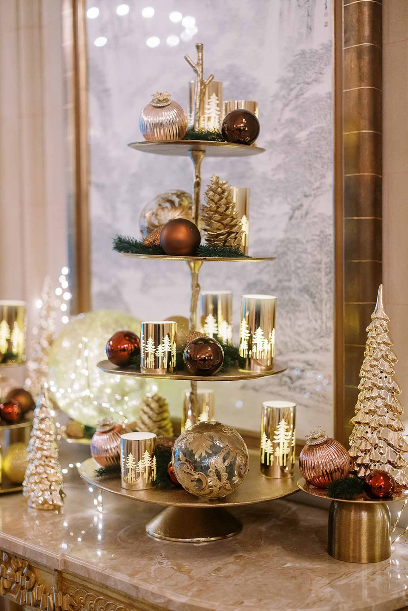 This image does not depict a wedding photograph. It shows a close-up decorative vignette of a four-tiered gold metal stand styled with Christmas/winter holiday ornaments, including deep red and chocolate brown glass baubles, a large silver filigree ornament, gold pine cones, lit gold cylindrical candle holders with white tree silhouette cutouts, and small evergreen sprigs. The display is set on a marble surface alongside ceramic gold Christmas trees and a glowing orb with fairy lights. The background features a framed decorative panel and additional warm fairy lighting. The overall decor palette is gold, deep red, chocolate brown, and copper in a classic winter holiday style. No people, wedding attire, floral arrangements, or wedding-related elements are present in this image.
