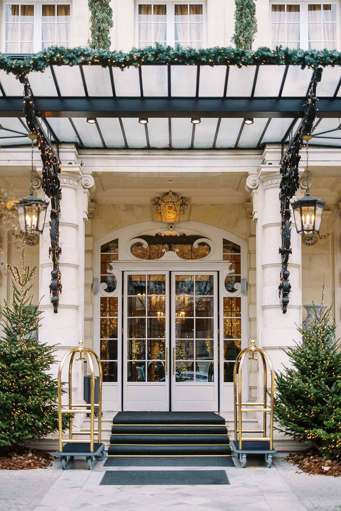 Exterior entrance shot of a grand Parisian hotel or private mansion, decorated for the winter holiday season. The facade features classical stone columns, ornate ironwork lanterns with warm candlelight bulbs, and a glass canopy draped with evergreen garland strung with warm white fairy lights. A gilded crest with the letter 'B' topped by a crown is mounted above the arched white double doors, which are flanked by undecorated Christmas trees wrapped in warm fairy lights. Two brass hotel luggage carts with black bases are positioned symmetrically on either side of the black-carpeted entrance steps. The warm golden light visible through the glass doors suggests an illuminated interior with additional festive lighting. Wide architectural shot with a straight-on, centered composition. Potential venue feature image.